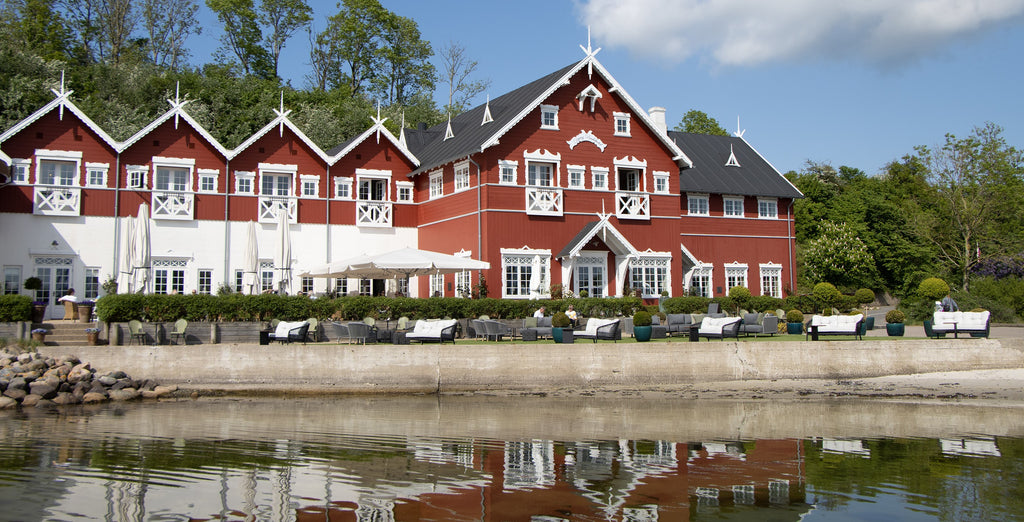 Dyvig Badehotel, red white hotel building in the nature with view over the sea. At the foreground there is a outdoor lounge area with Cane-line lounge sofas
