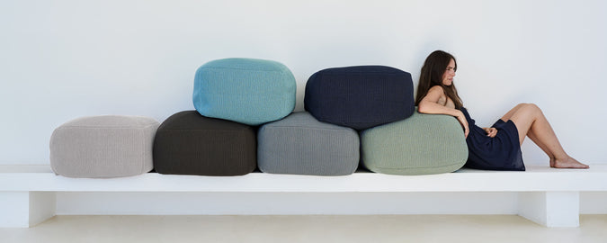 A woman relaxes beside a colorful arrangement of cushions in various shapes and shades, creating a visually appealing composition.