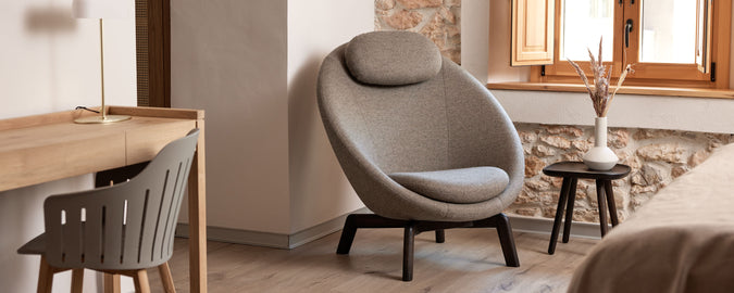 Gemütlicher Sitzbereich mit einem modernen Stuhl mit abgerundetem Design, einem kleinen Beistelltisch und einem minimalistischen Schreibtisch. Natürliches Licht strömt durch ein Fenster.