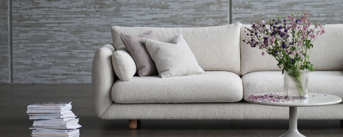 Stylische hellfarbene Couch mit dekorativen Kissen, begleitet von einem kleinen Tisch mit einer Vase voller Blumen und einem Stapel Zeitschriften.