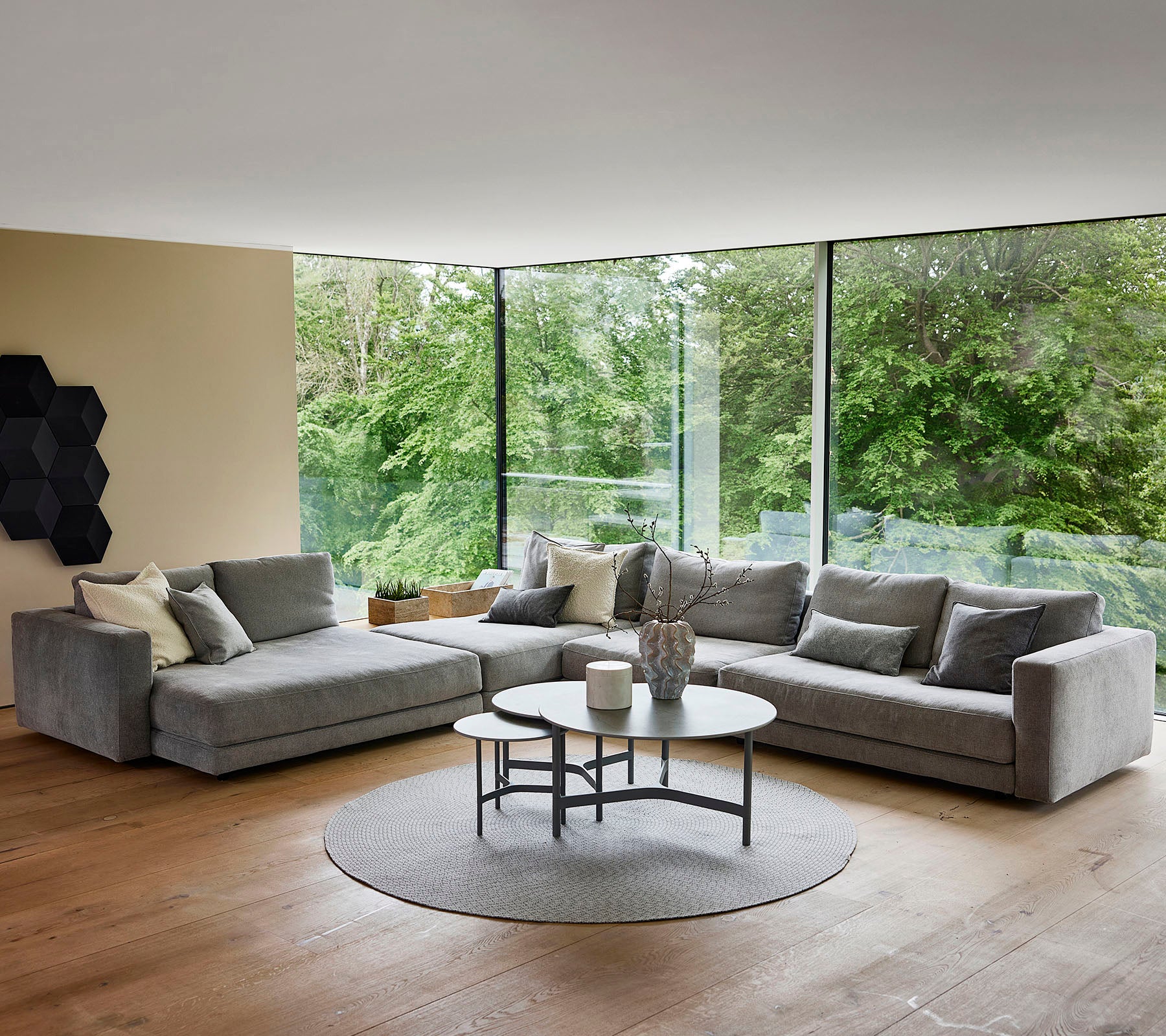 Ein modernes Wohnzimmer mit einem grauen Ecksofa mit dekorativen Kissen, einem runden Couchtisch und einem runden Teppich, umgeben von Grünpflanzen.
