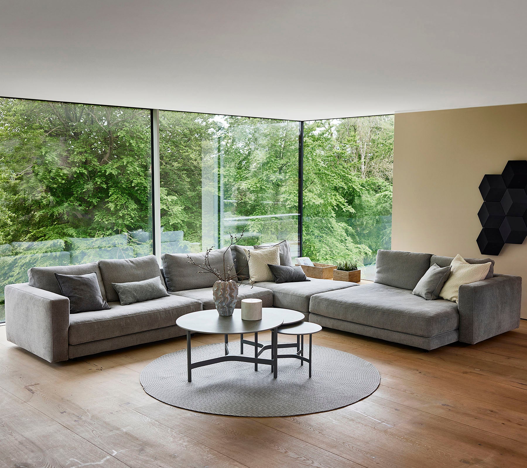 Ein stilvolles, modernes Wohnzimmer mit einem bequemen grauen Ecksofa, einem runden Couchtisch und großen Fenstern, die die grüne Umgebung draußen zeigen.