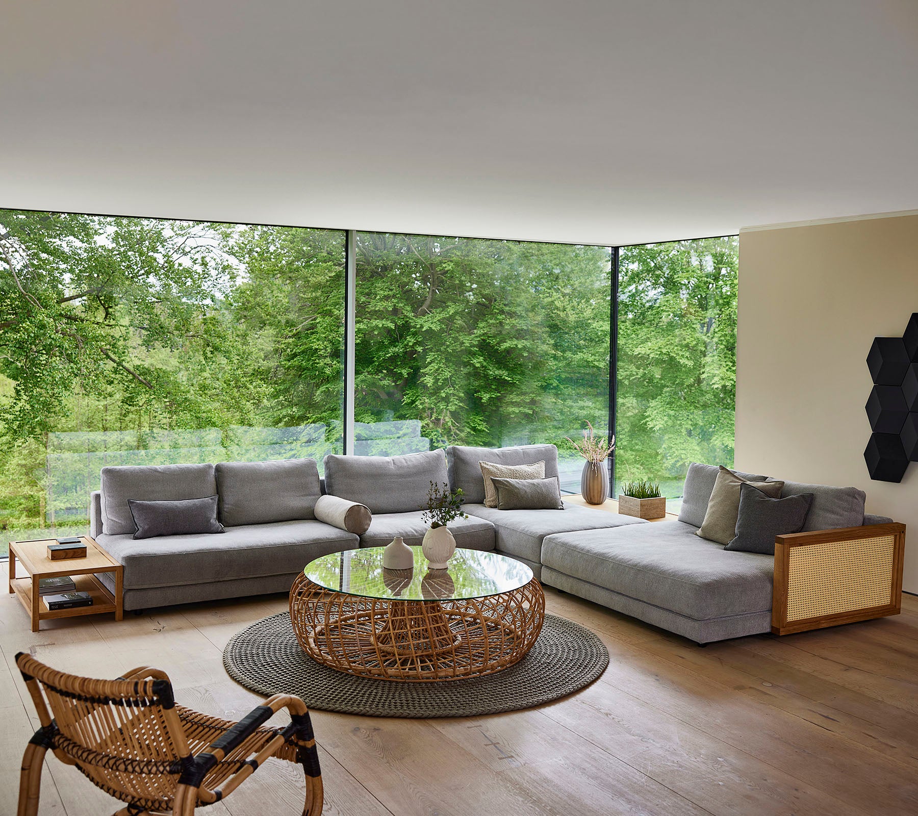 Ein modernes Sitzelement mit einem grauen sectional Sofa, einem runden Couchtisch und einem geflochtenen Stuhl, umgeben von Grün.