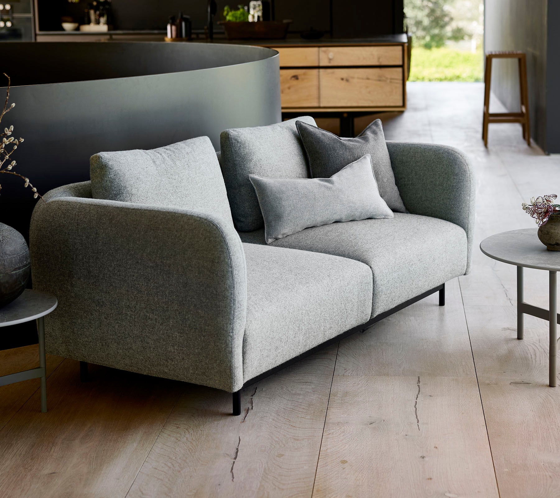 Modernes graues Sofa mit plüschigen Kissen, kombiniert mit einem kleinen runden Tisch, vor einem minimalistischen Hintergrund.