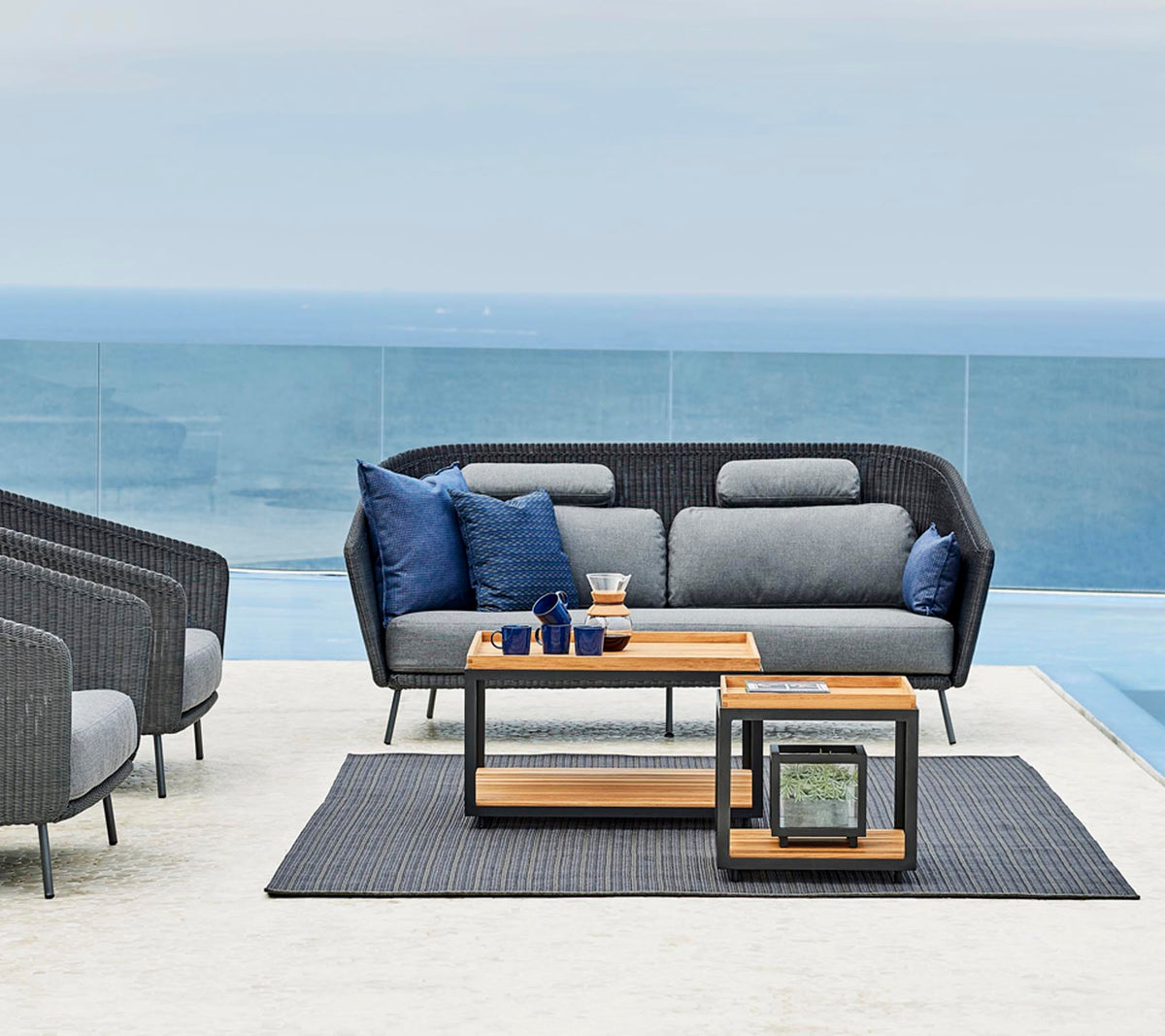 Stylische Außensitzanordnung mit einem grauen Sofa mit blauen Kissen, einem Holztisch und einem passenden Teppich.