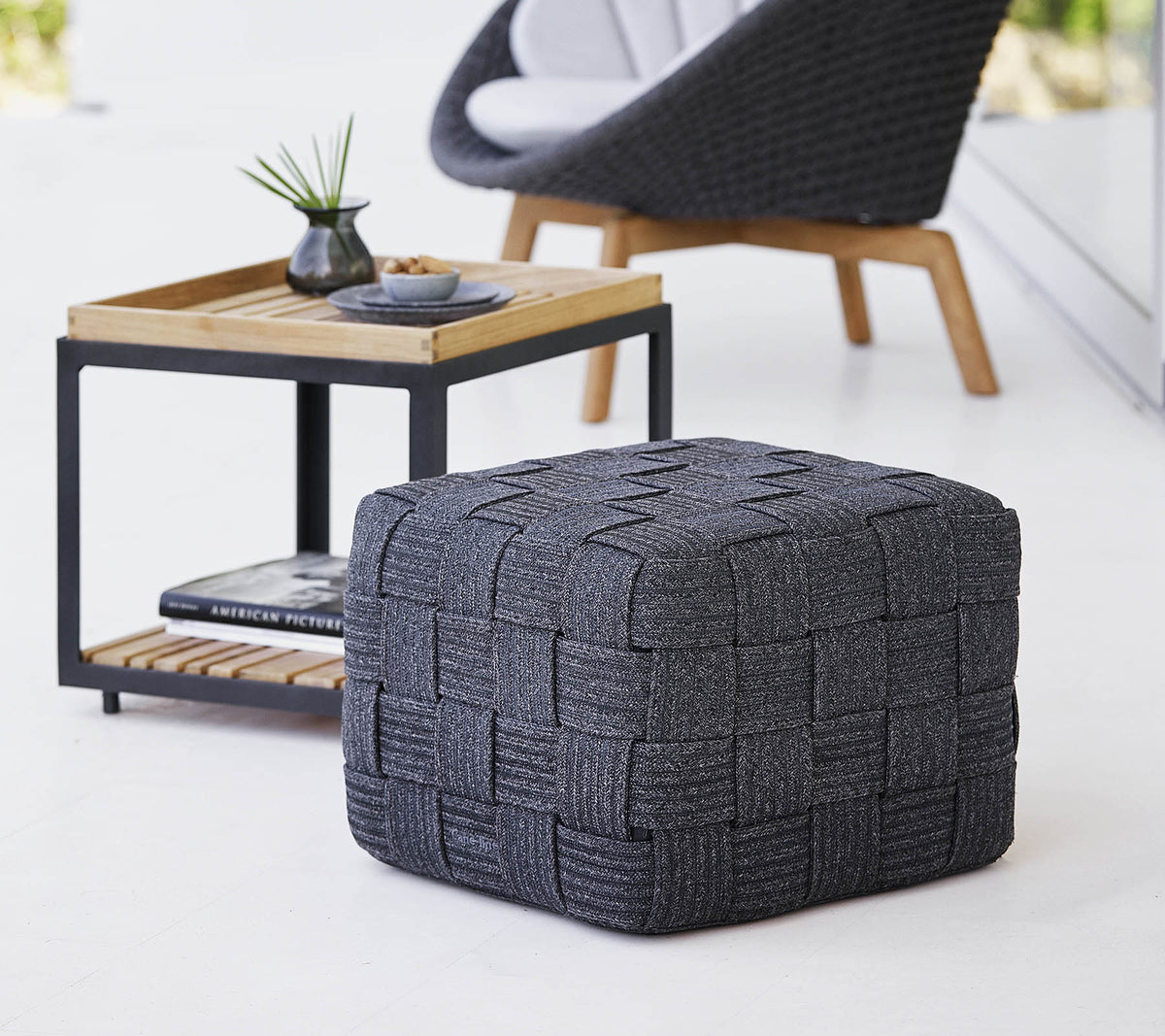 Ein strukturierter grauer gewebter Pouf, der neben einem minimalistischen Holztisch steht, der kleine Dekorationsartikel enthält.