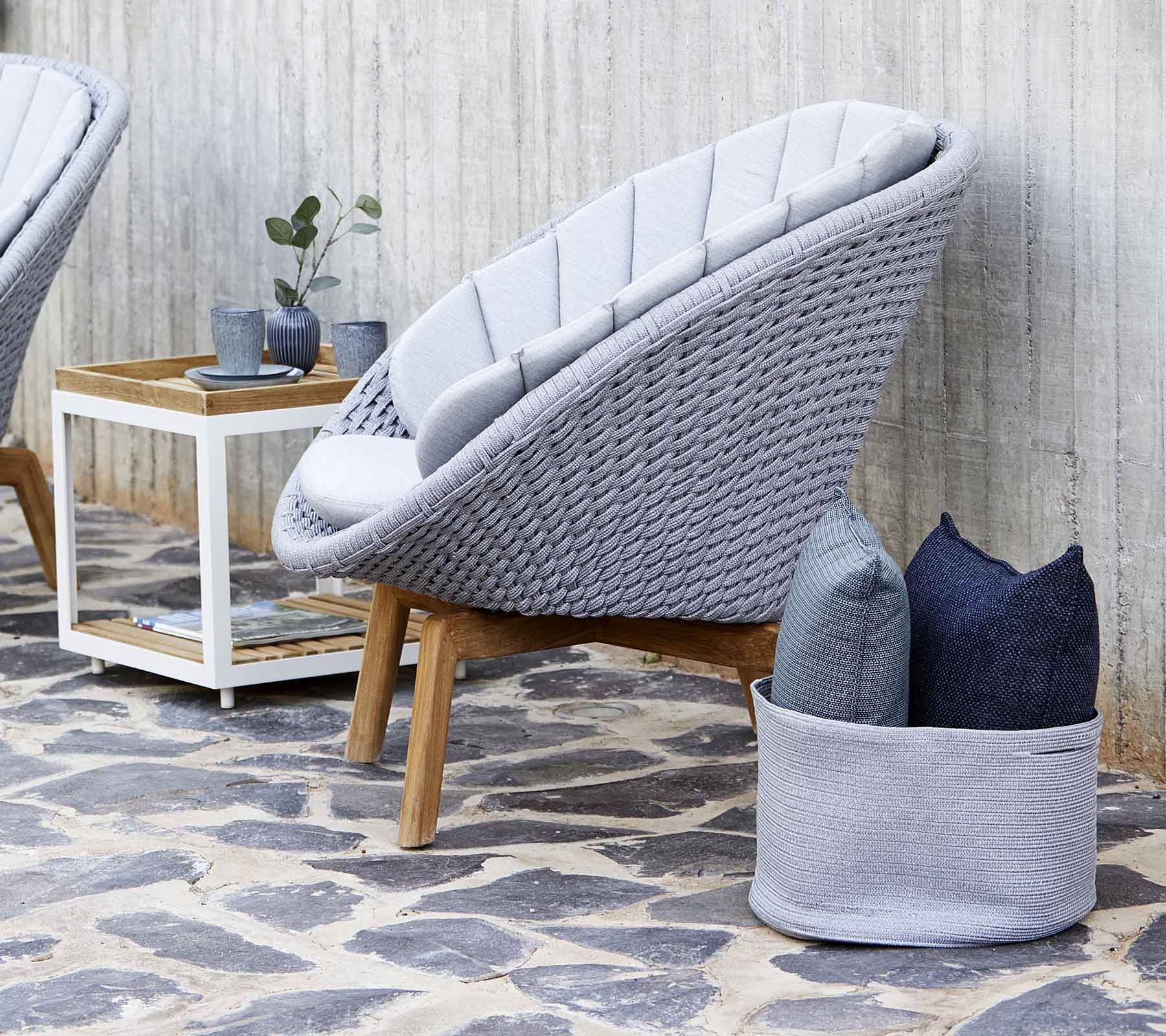 Moderne graue Lounge-Stuhl mit einem gewebten Design, begleitet von Kissen und einem kleinen Holzbeistelltisch. Der Steinboden verbessert die Ästhetik.