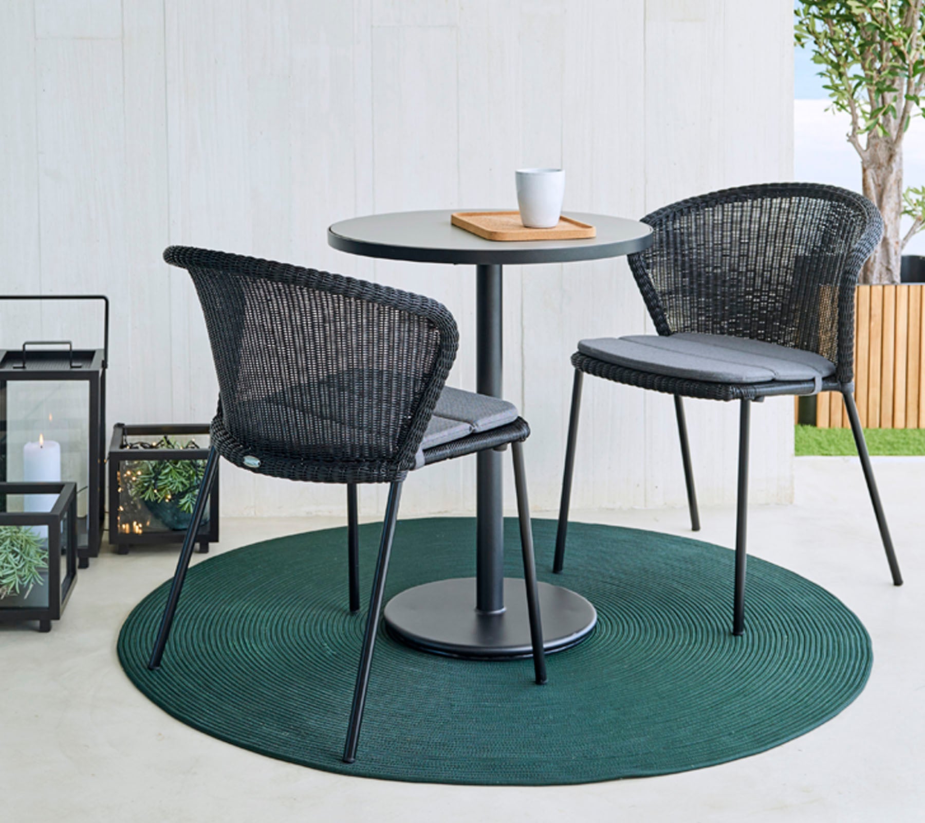 Ein runder Tisch mit zwei schwarzen geflochtenen Stühlen auf einem grünen Teppich, begleitet von einer Kaffeetasse und einem Holzbrett. Dekorative Laternen im Hintergrund.