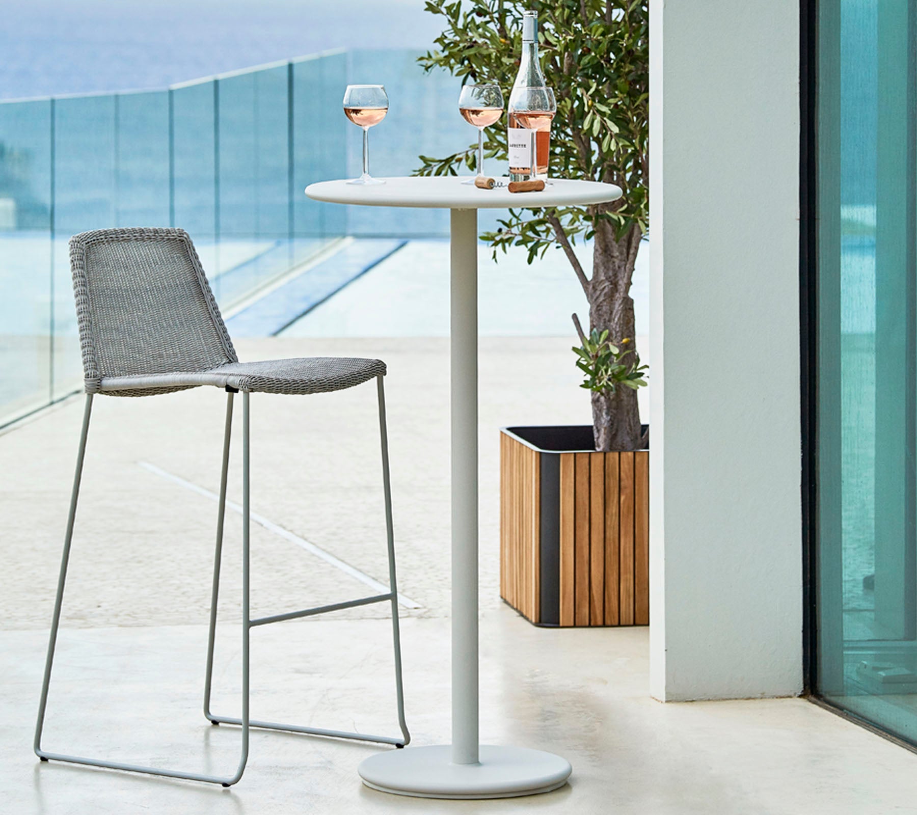 Stilvolles Outdoor-Setting mit einem hohen Tisch, einem grauen Barhocker und zwei Gläsern Rosé, die mit einer Flasche auf dem Tisch kombiniert sind.