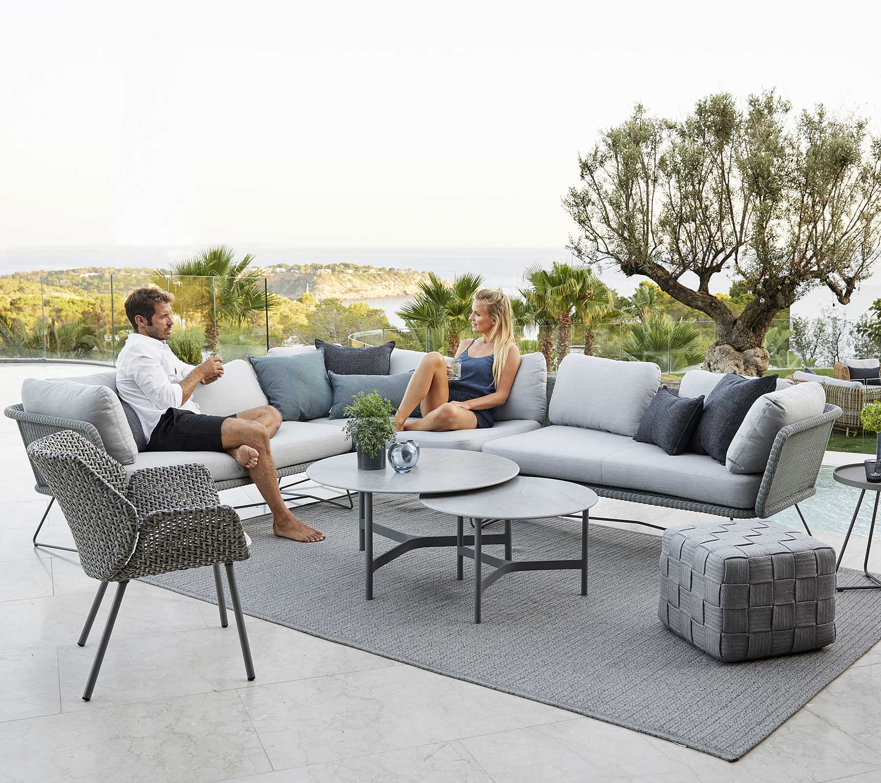 Ein modernes Outdoor-Sitzbereich mit einem Sofa, einem Sessel und einem Couchtisch, in dem zwei Personen entspannen, umgeben von Grün und einer malerischen Aussicht.
