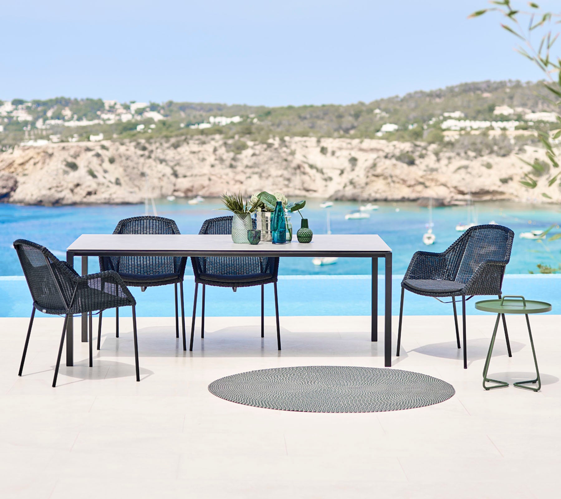 Ein modernes Outdoor-Esszimmer-Set mit einem rechteckigen Tisch, geflochtenen Stühlen und einem kleinen Beistelltisch, mit einer malerischen Strandkulisse.