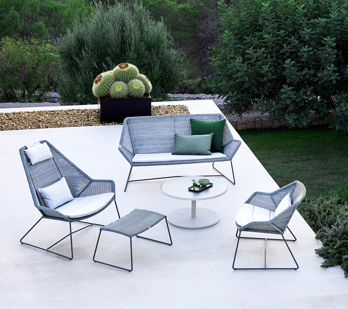 Stylisches Außen-Sitzarrangement mit einem Sofa, zwei Armlehnstühlen, einem Fußhocker und einem Couchtisch, ergänzt durch Grünpflanzen im Hintergrund.