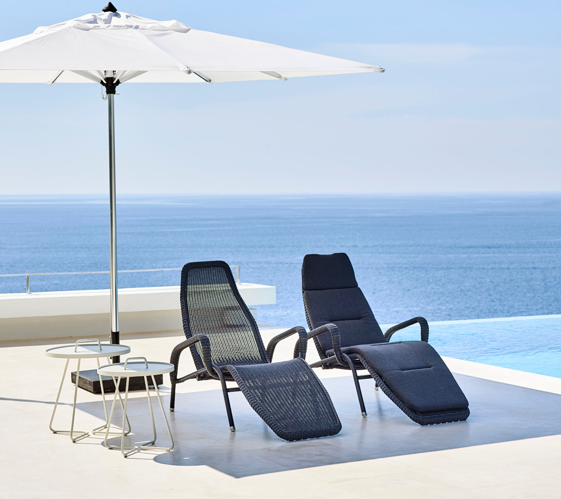 Zwei stilvolle Lounge-Stühle und ein kleiner Tisch stehen unter einem großen Sonnenschirm, mit einem ruhigen blauen Meer im Hintergrund.