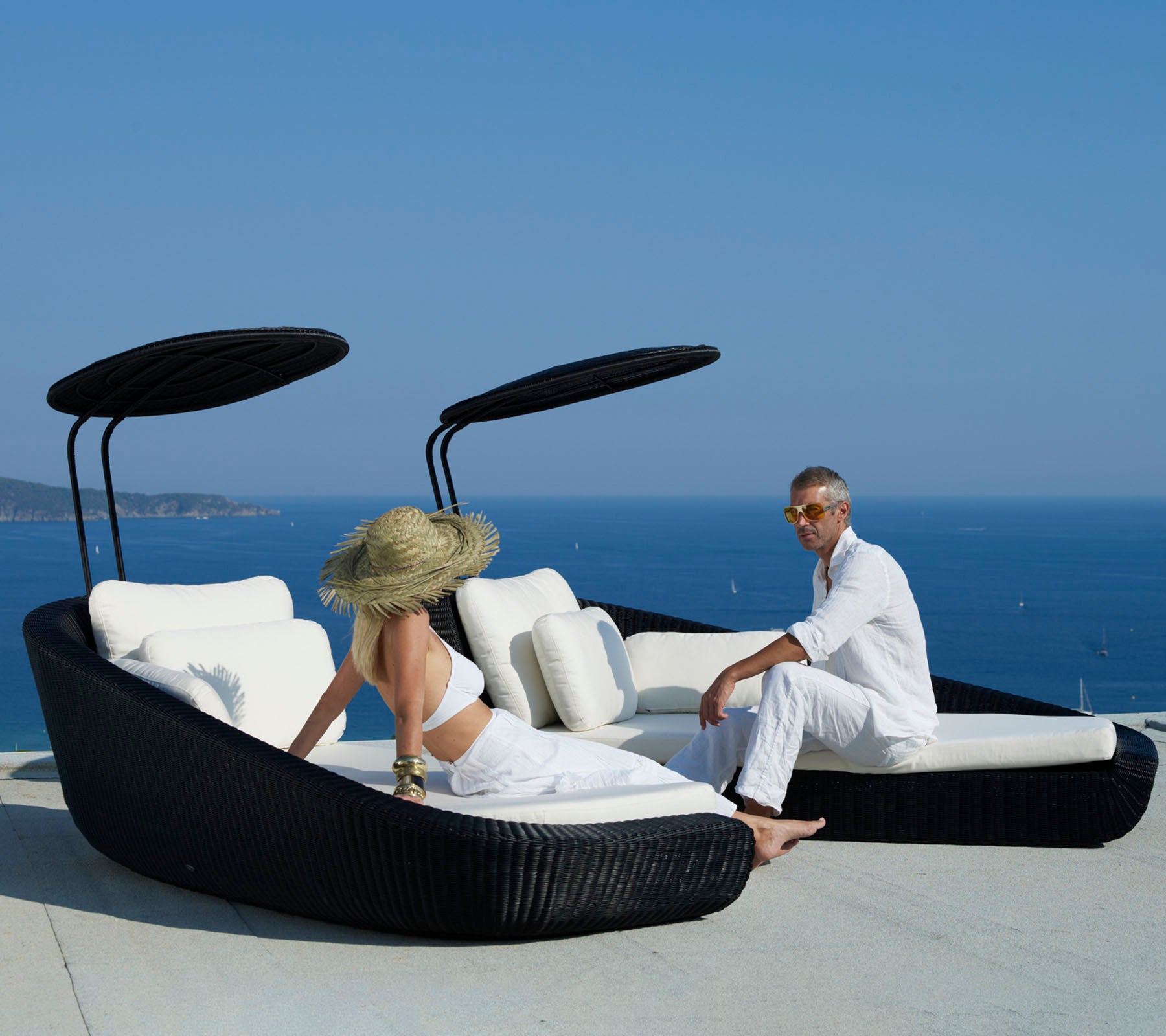 Zwei Personen entspannen auf einer stilvollen, geschwungenen Sonnenliege mit Blick auf das malerische Meer, ausgestattet mit einem eleganten schwarzen Design und flauschigen weißen Kissen.