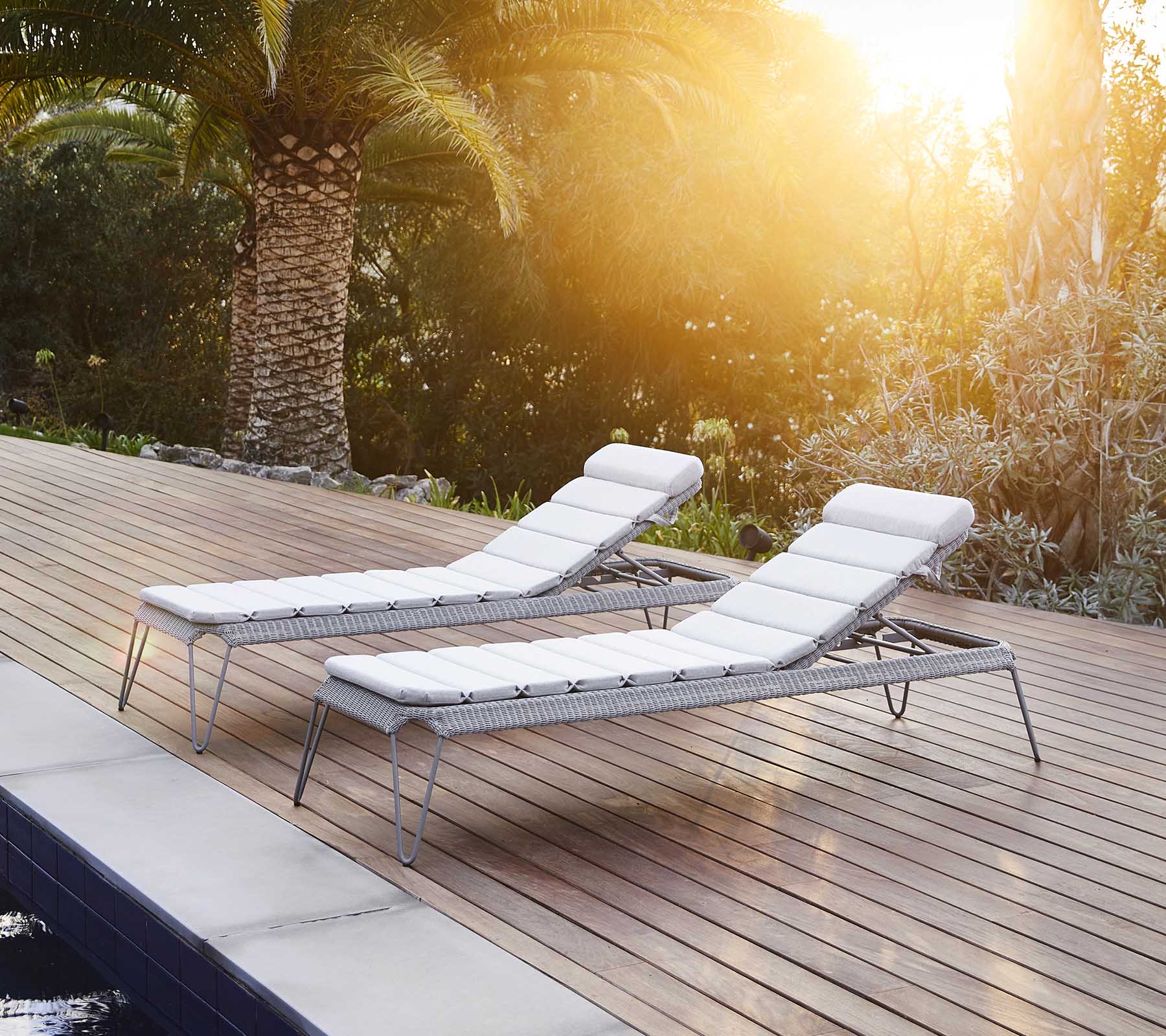 Zwei stilvolle Loungestühle mit weißen Kissen, neben einem Pool positioniert und von üppigem Grün umgeben, die sich im warmen Sonnenlicht aalen.