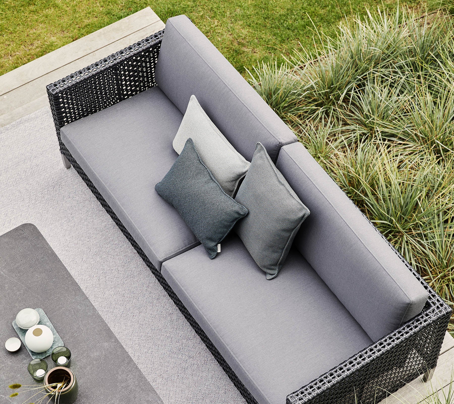 Stilvolles graues Sofa mit dekorativen Kissen, umgeben von üppigem Grün und modernen dekorativen Elementen. Perfekt zum Entspannen und für Zusammenkünfte.