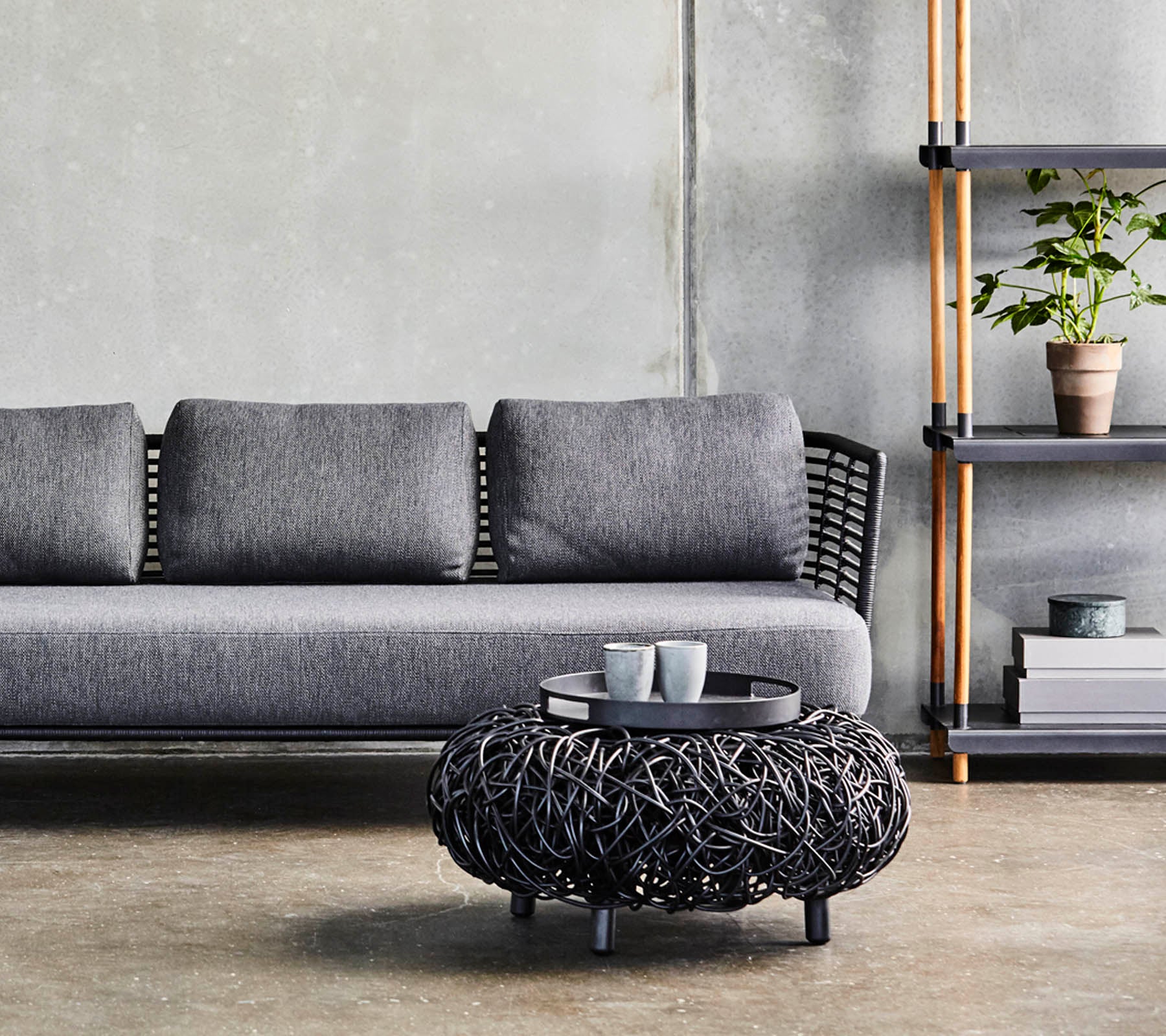 Ein modernes graues Sofa mit Kissen, kombiniert mit einem runden schwarzen geflochtenen Couchtisch und einem minimalistischen Regal, das Pflanzen und Dekoration zeigt.