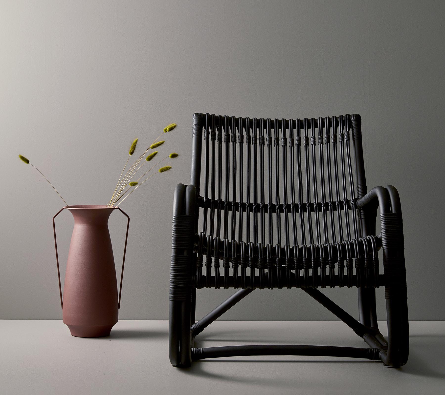 Stylischer schwarzer geflochtener Stuhl neben einer rosa Vase mit grünen Stängeln, vor einem minimalistischen grauen Hintergrund.