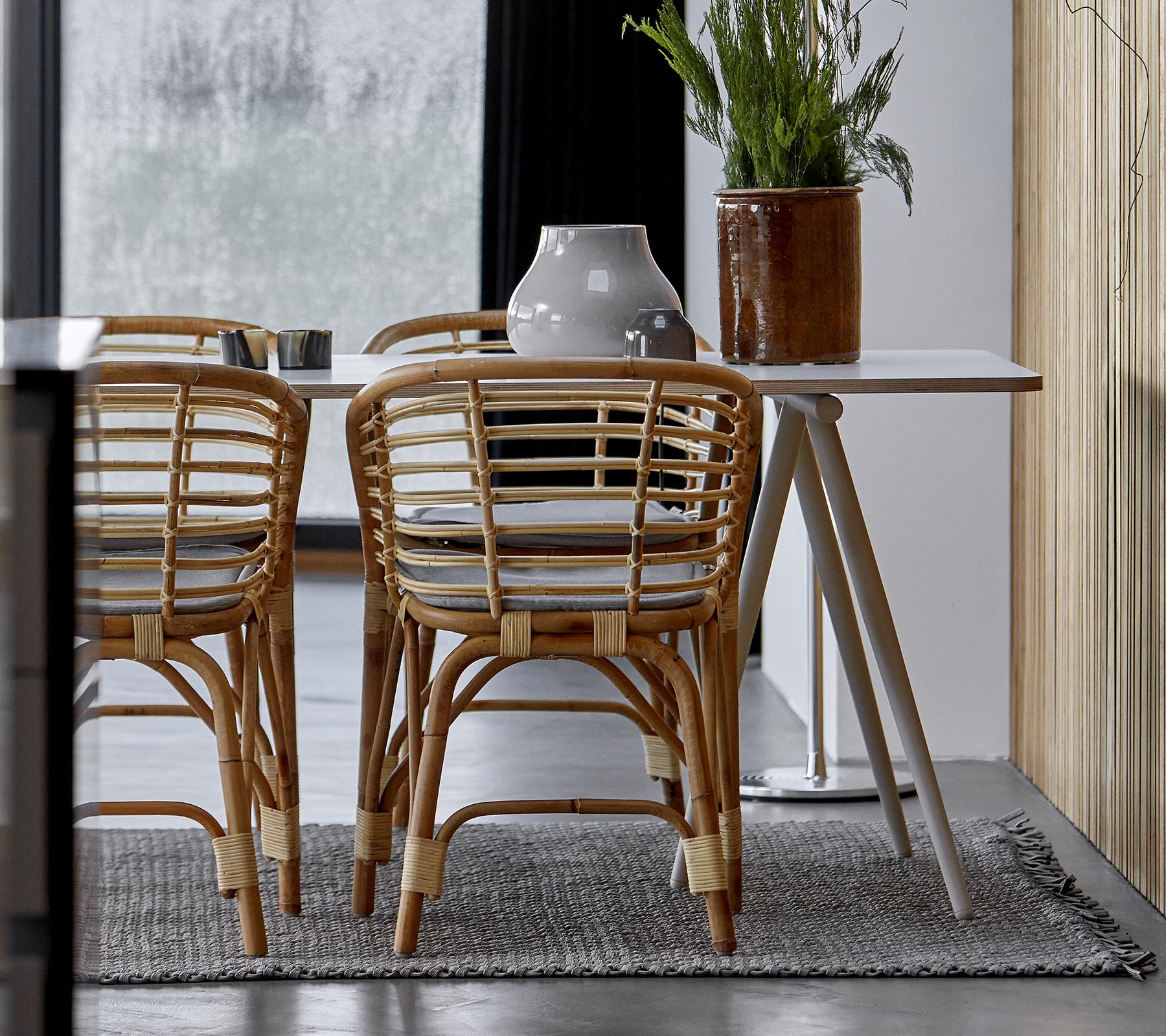 Ein modernes Esszimmer-Setup mit Rattanstühlen, einem eleganten Tisch und dekorativem Grün in einem stilvollen Innenraum.
