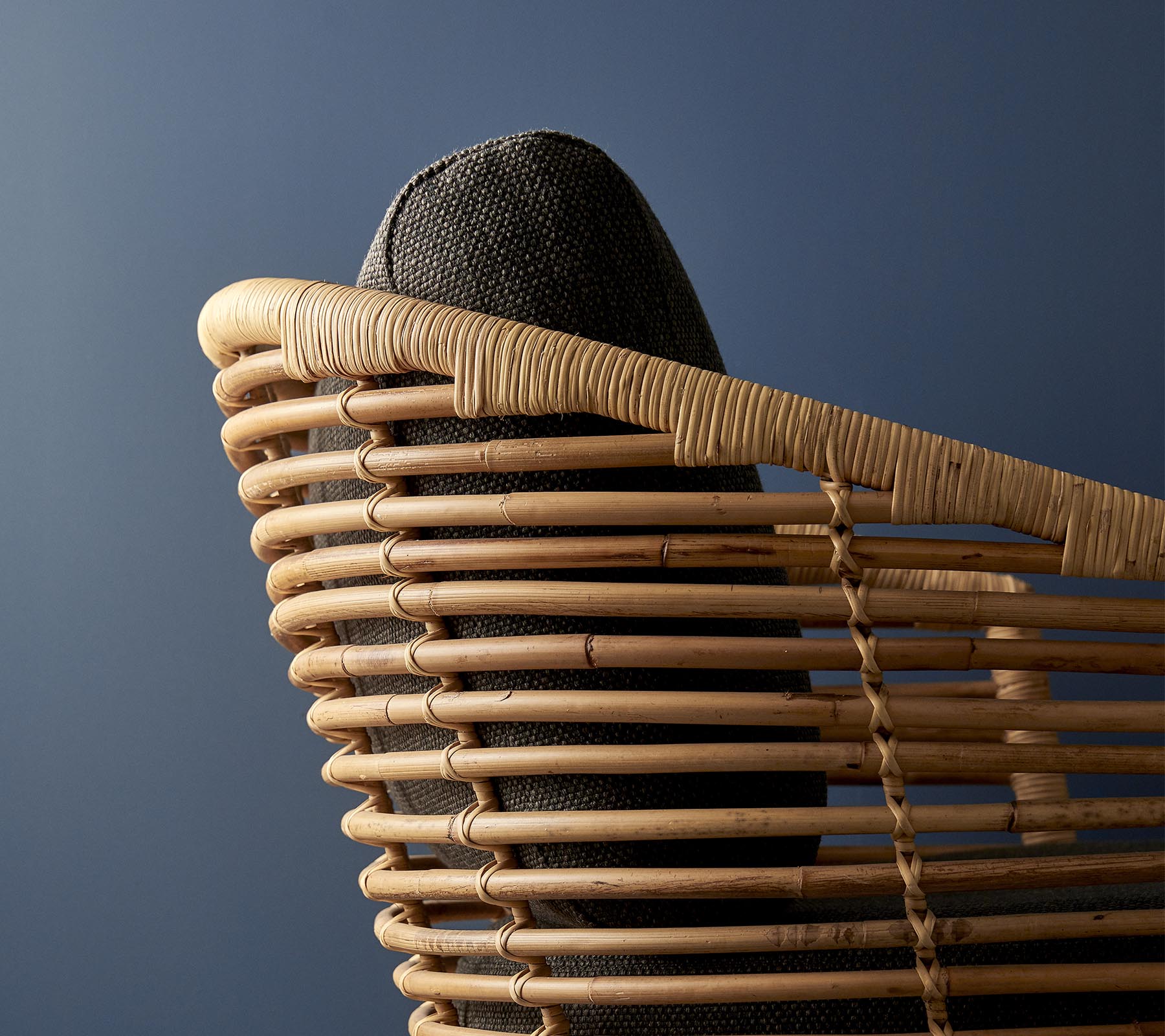 Stilvoller gewebter Stuhl mit einer geschwungenen Rückenlehne und dunklem gepolstertem Sitz vor einem gedämpften blauen Hintergrund.