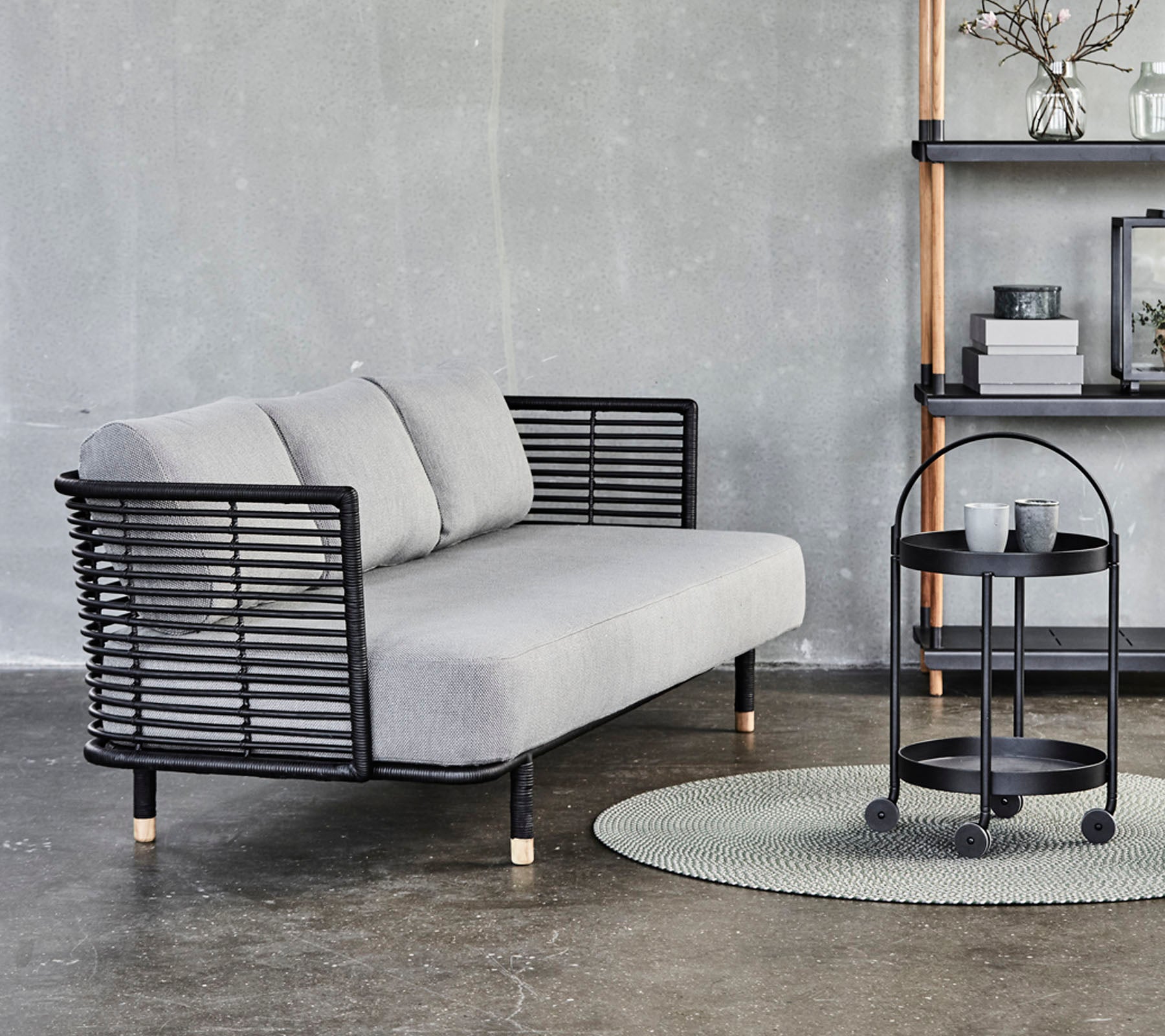 Elegantes graues Sofa mit schwarzen Metallakzenten und Kissen, kombiniert mit einem runden Beistelltisch auf einem strukturierten Teppich vor einem neutralen Hintergrund.