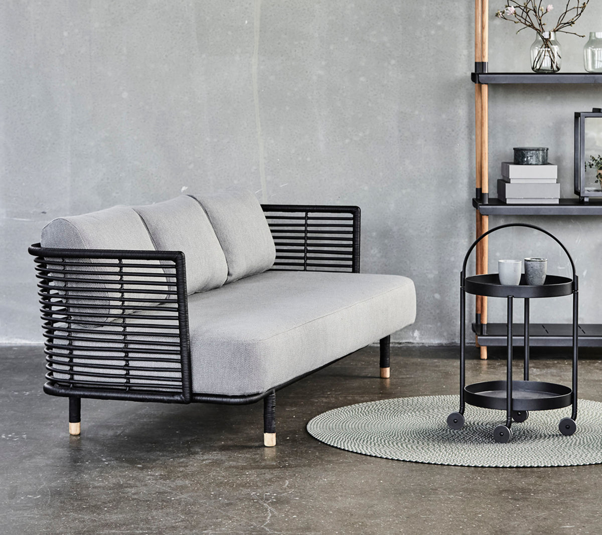 Elegantes graues Sofa mit schwarzen Metallakzenten und Kissen, kombiniert mit einem runden Beistelltisch auf einem strukturierten Teppich vor einem neutralen Hintergrund.