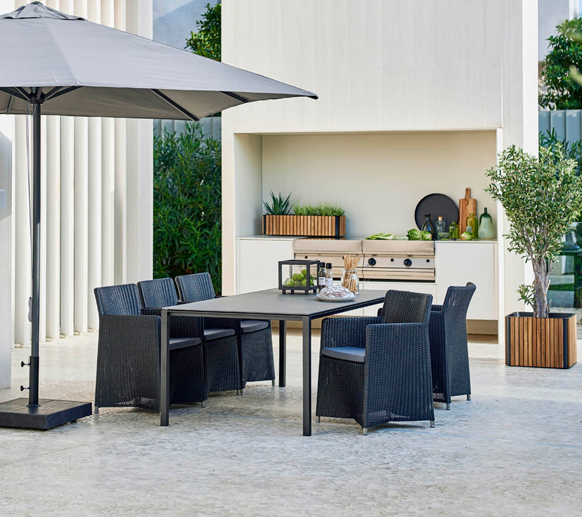 Modernes Outdoor-Esstischset mit einem großen Tisch, sechs Stühlen, einem stilvollen Sonnenschirm und einem Hintergrund mit Grün und dekorativen Elementen.