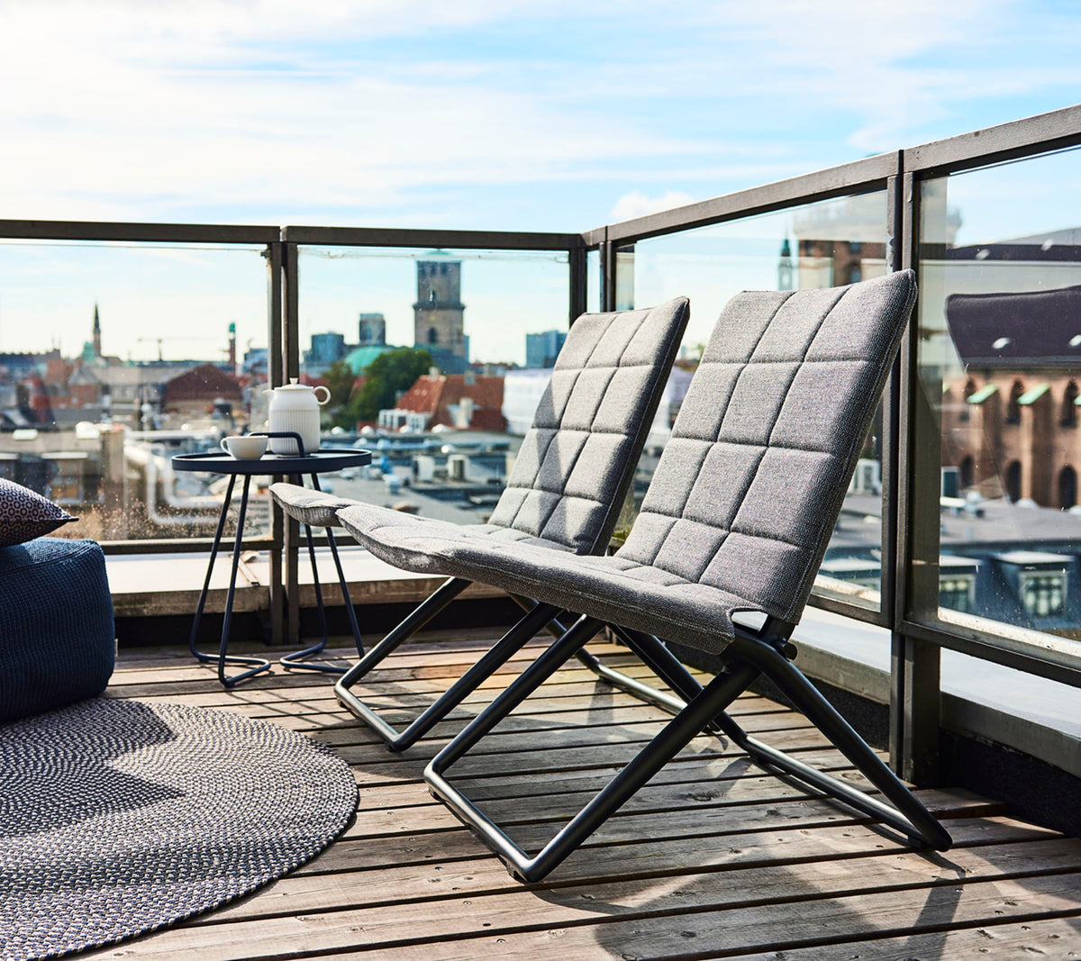 Zwei moderne Lounge-Stühle mit schlanken Rahmen und strukturierten Kissen, begleitet von einem kleinen Beistelltisch und Ausblick auf die Stadt im Hintergrund.