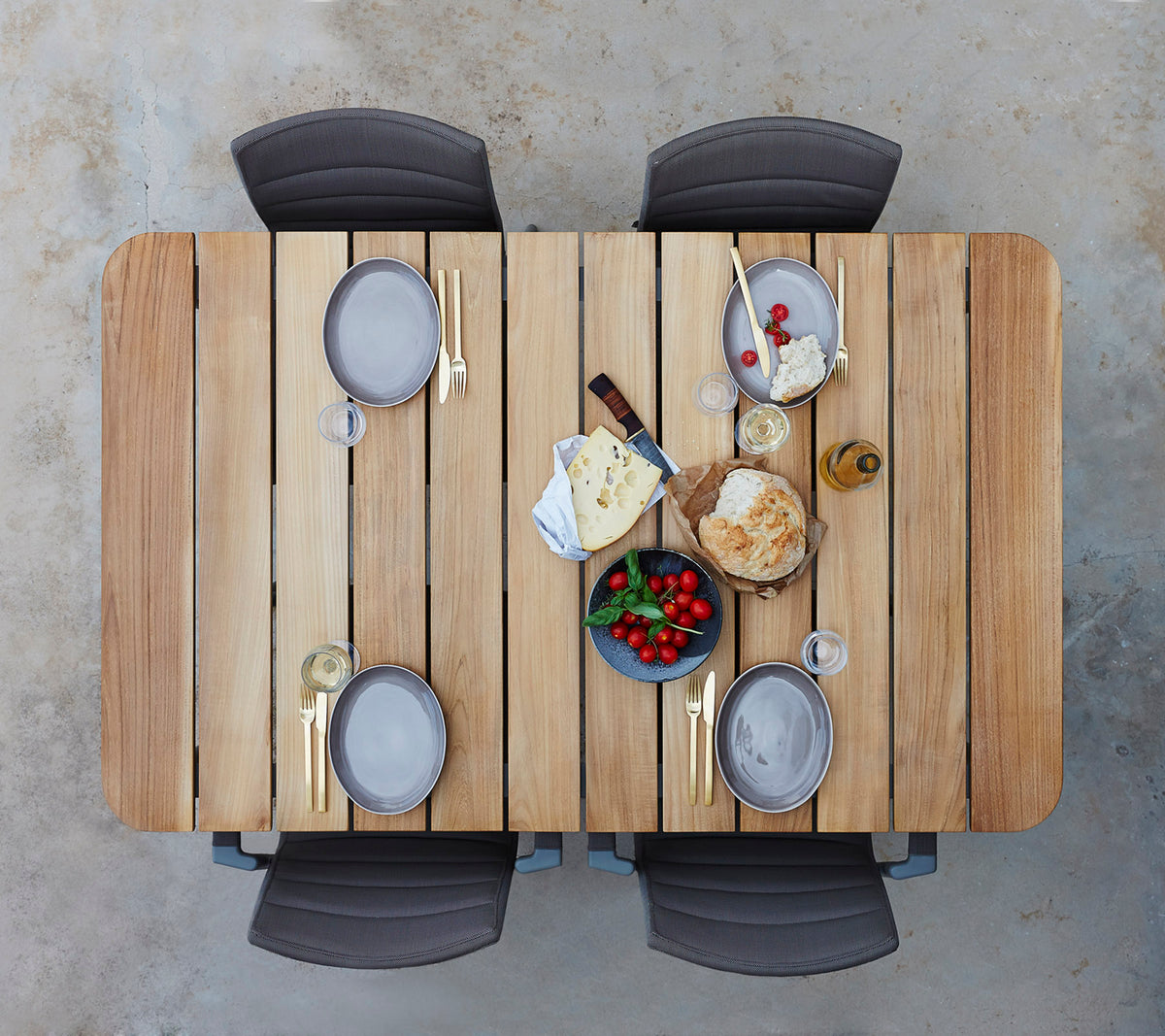 Ein Holztisch, gedeckt mit Tellern, Gläsern und einer Auswahl an Käse, Obst und Brot, umgeben von vier Stühlen.