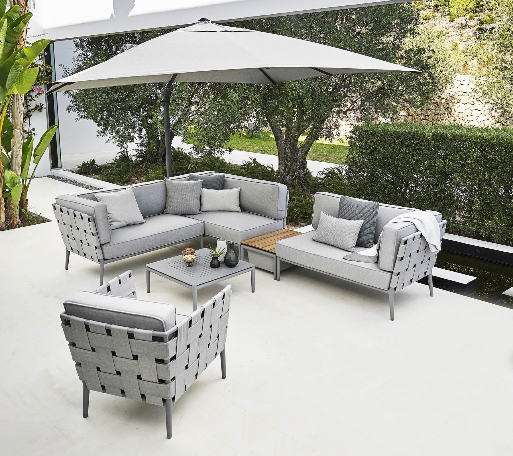 Stylische Outdoor-Sitzgelegenheit mit einem grauen Ecksofa, einem Sessel und einem Couchtisch unter einem großen Sonnenschirm.