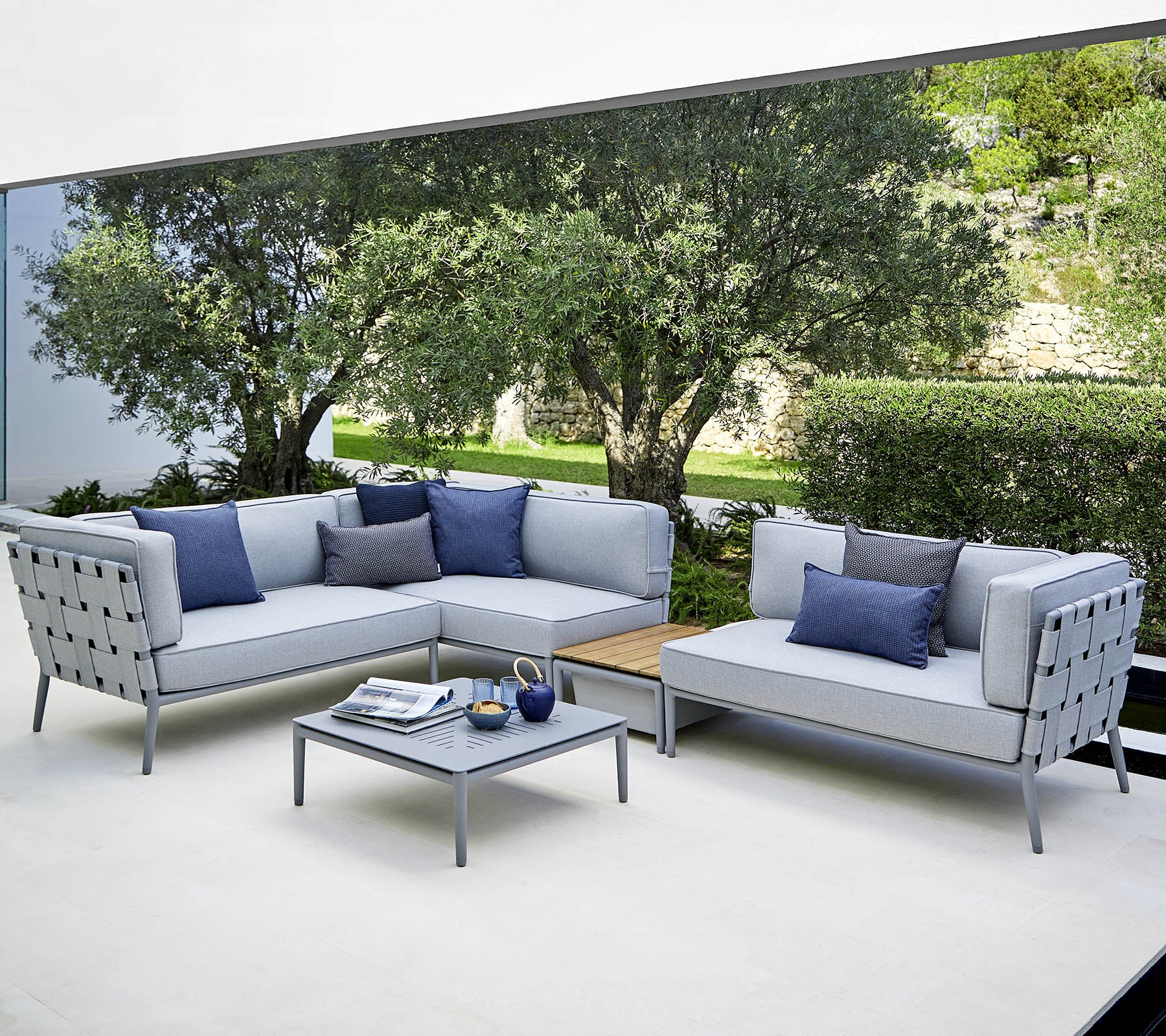 Eine moderne Außensitzanordnung mit hellgrauen Sofas und blauen Kissen sowie einem passenden Couchtisch, vor einem üppigen Hintergrund.