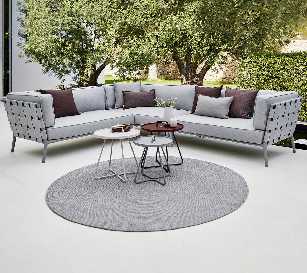Moderne Ecksofa in Hellgrau mit dekorativen Kissen, begleitet von stilvollen Couchtischen auf einem runden Teppich. Üppiges Grün im Hintergrund.