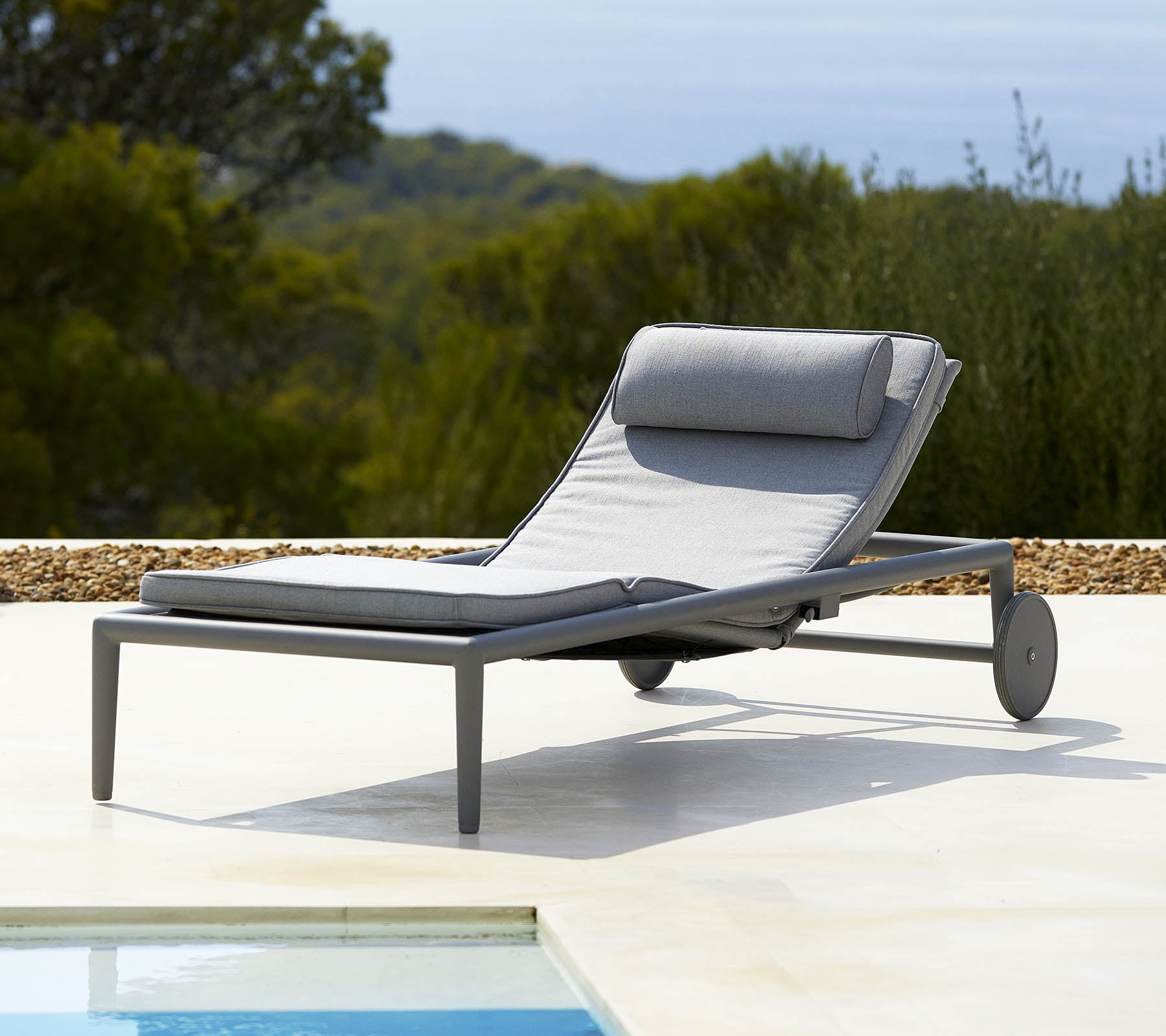 Bequemer Loungestuhl mit einem grauen Kissen und Rückenlehne, ausgestattet mit Rädern für einfache Mobilität, neben einem Pool und üppigem Grün im Hintergrund.