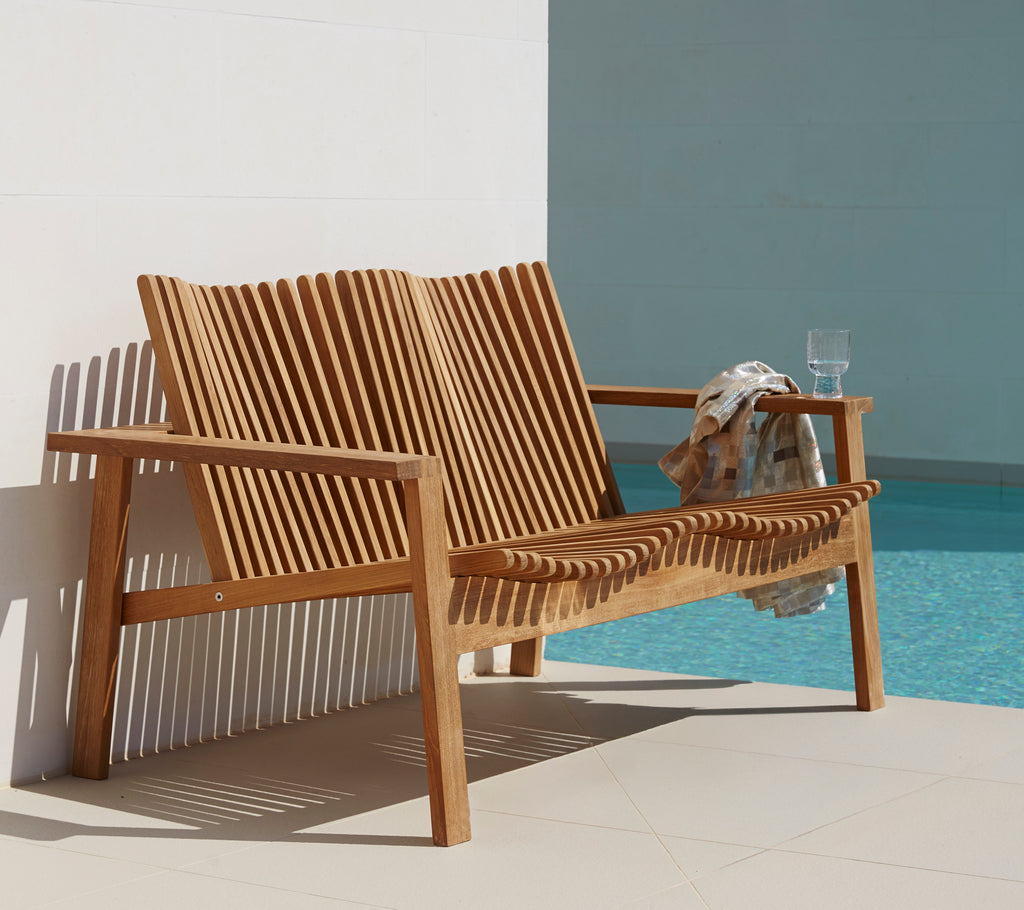 Seitenansicht eines eleganten, hölzernen 2-Sitzer Sofas mit Lamellen-Design, begleitet von einem Getränk und einem Handtuch. Ideal für entspanntes Sitzen.