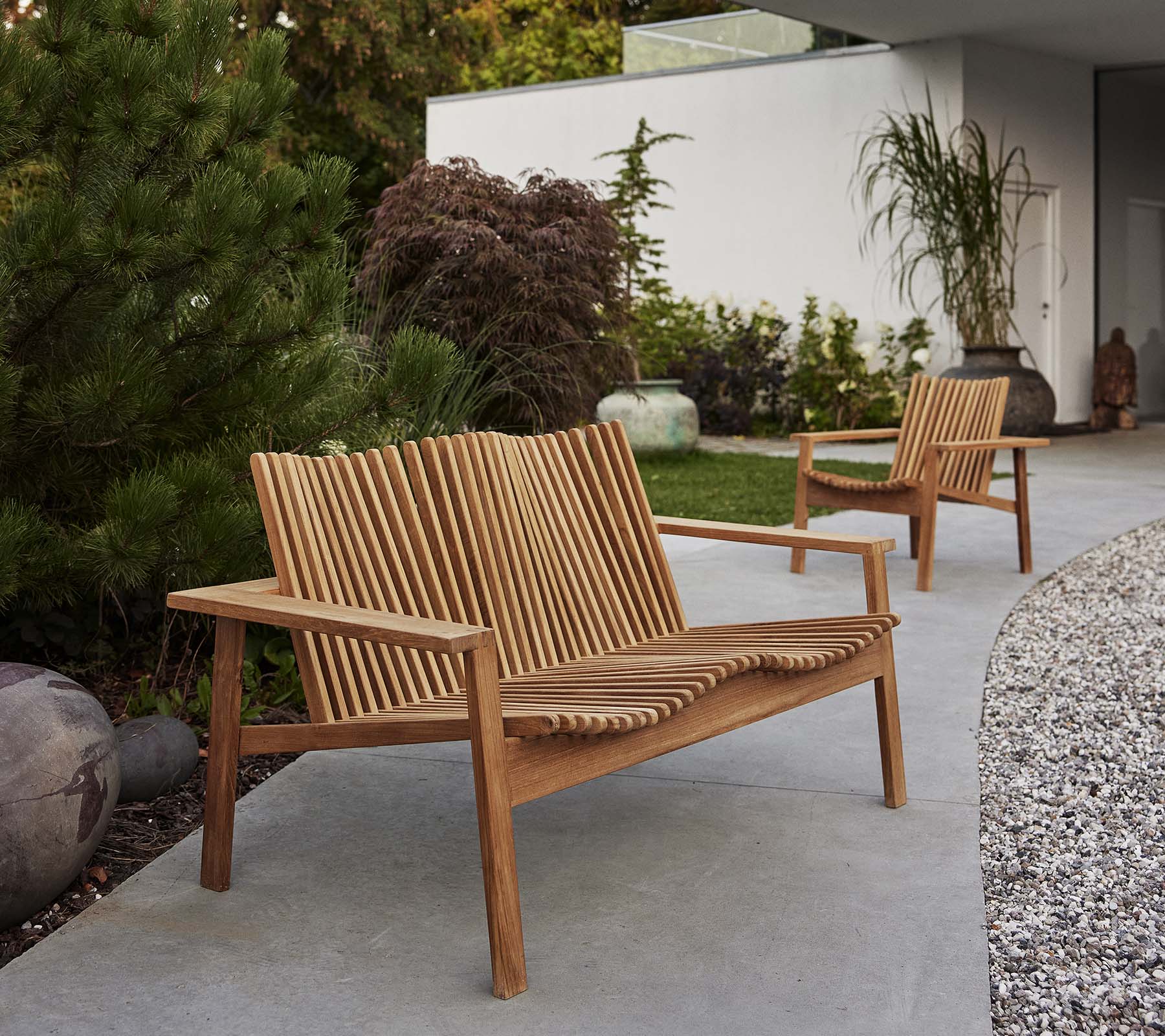 Zwei hölzerne Amaze-Loungestühle und ein passendes Sofa verfügen über ein Latten-Design, umgeben von Pflanzen und dekorativen Elementen.