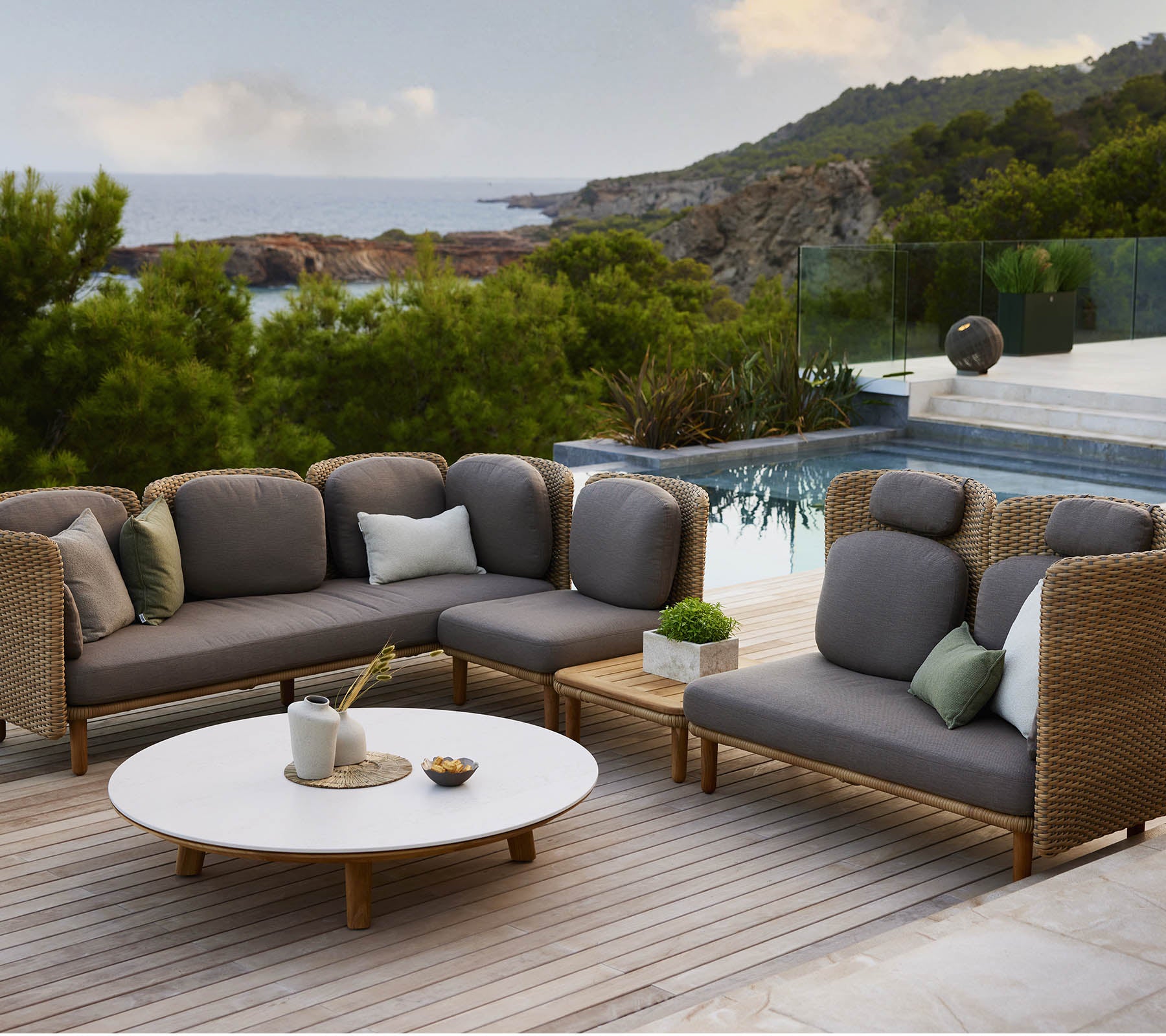 Stylische Loungemöbel mit grauen Kissen und dekorativen Kissen, begleitet von einem runden Tisch, vor einer malerischen Kulisse.
