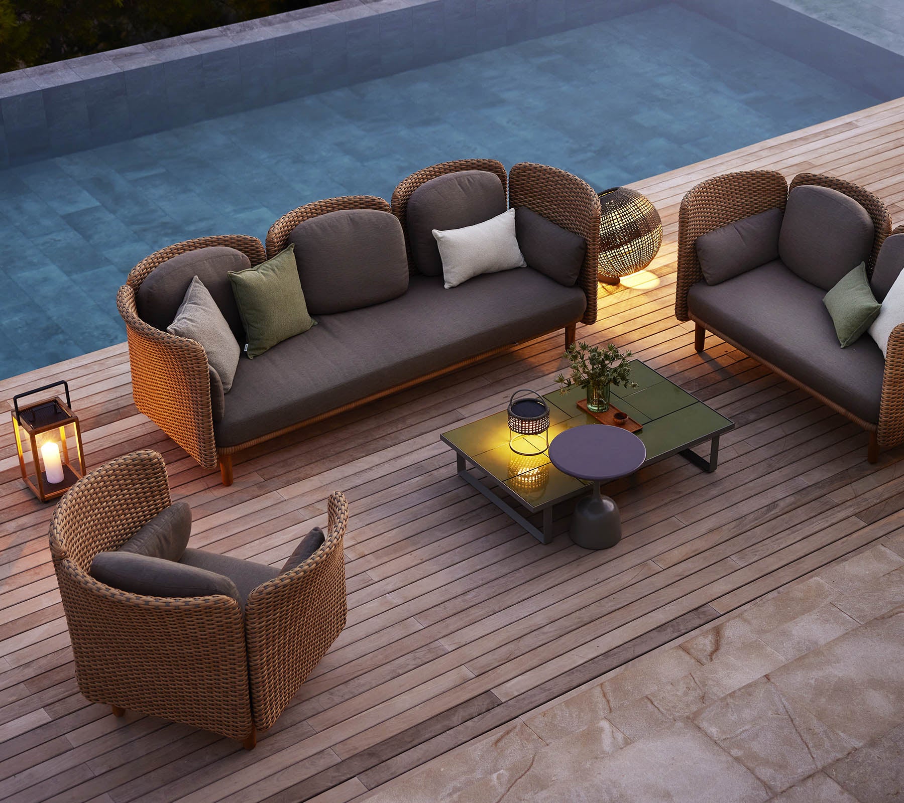 Eine gemütliche Sitzgelegenheit mit einem Sofa und Stühlen mit Kissen, umgeben von Grün und einem Pool, auf einer Holzterrasse.