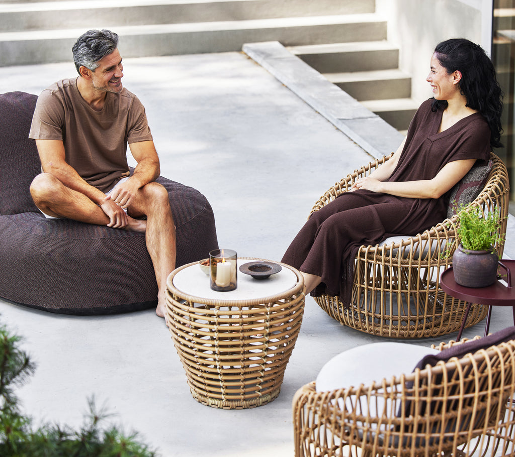 Zwei Personen sitzen zusammen, eine auf einem gemütlichen Sessel und die andere auf einem runden Korbstuhl, mit einem kleinen Korbtisch zwischen ihnen.