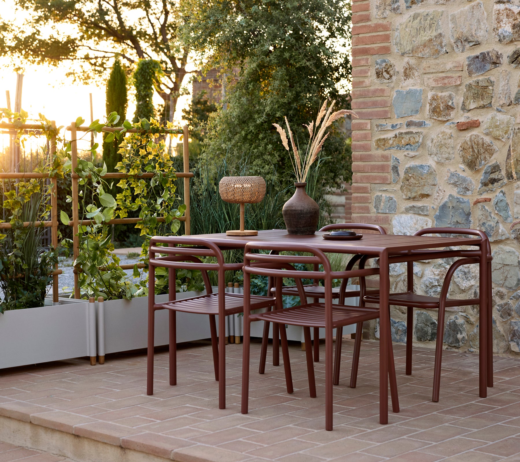 Außenbereich mit einem Bliss-Tisch mit Stühlen, einem Bliss-Sessel und einem Sticks-Blumenkübel, umgeben von Grün bei Sonnenuntergang.