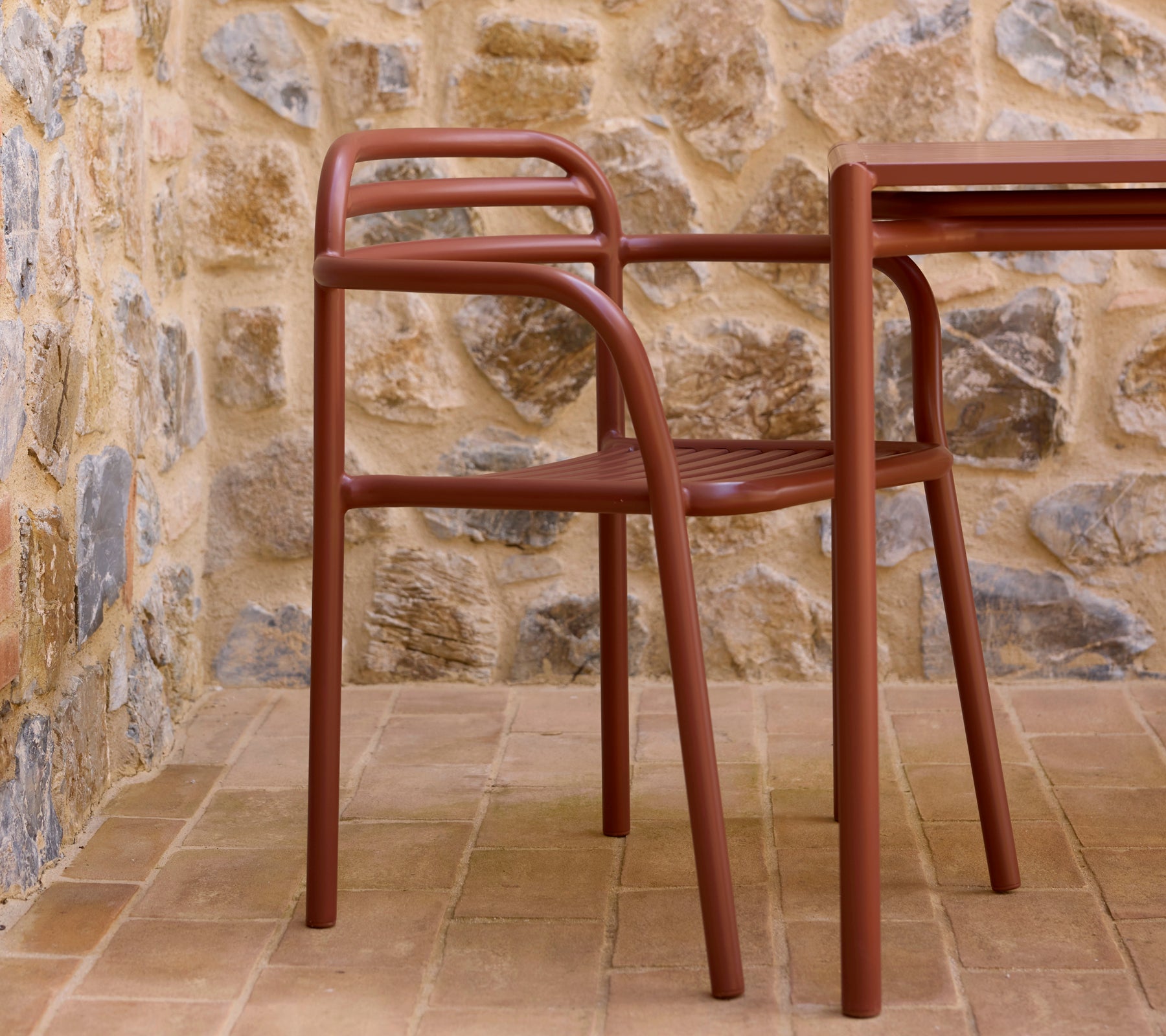 Wüstenroter Bliss-Sessel neben einem passenden Tisch, an einer strukturierten Steinmauer platziert. Eine stilvolle und moderne Sitzanordnung.