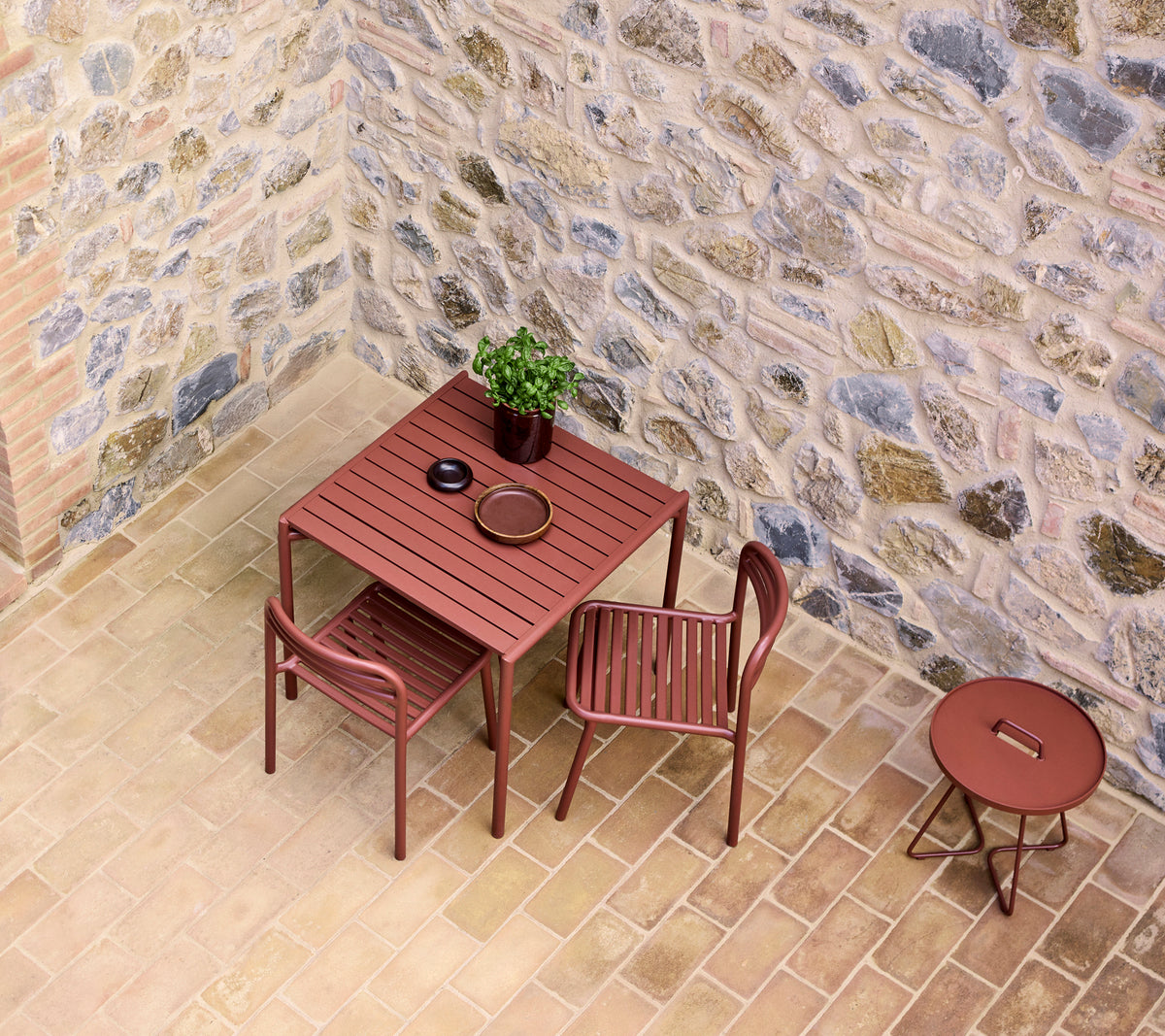 ein roter Café-Tisch mit zwei Stühlen, einem kleinen Pflanze und einem Teller, vor einer Steinmauer.