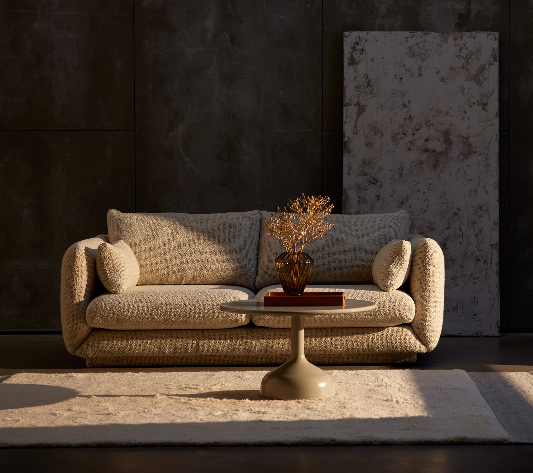 Bloom 2-Sitzer Sofa in Sand Flair mit einem Glaze-Couchtisch und einer dekorativen Pflanze auf einem Teppich, vor einer strukturierten dunklen Wand.