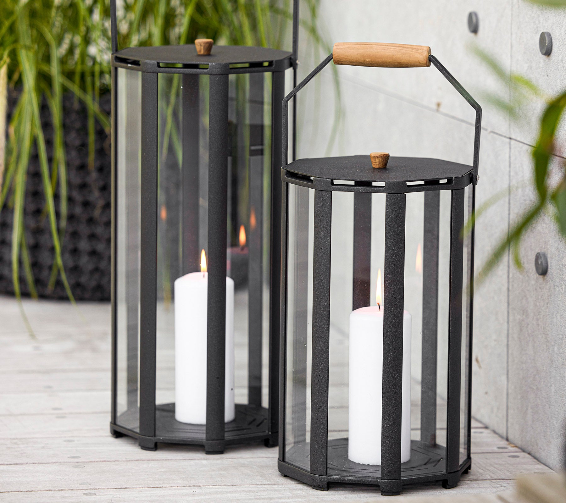 Zwei stilvolle Laternen mit modernem Design, ausgestattet mit Glaspaneelen und einem Holzgriff, die weiße Kerzen im Inneren halten.