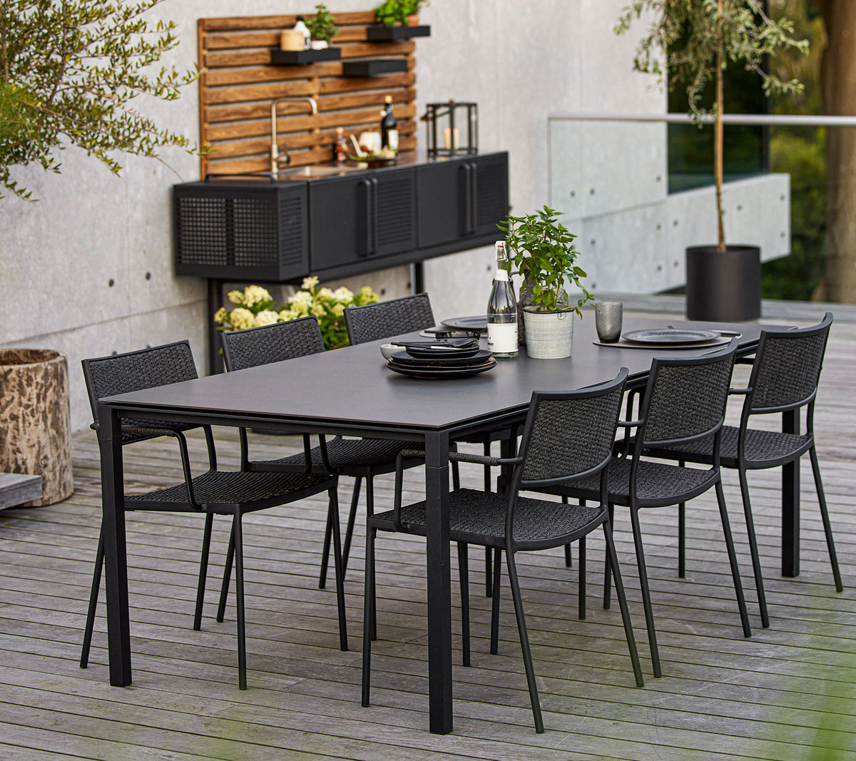 Ein modernes Esszimmer-Set mit einem großen rechteckigen Tisch und sechs schwarzen geflochtenen Stühlen, akzentuiert durch dekorative Elemente und Grünpflanzen.