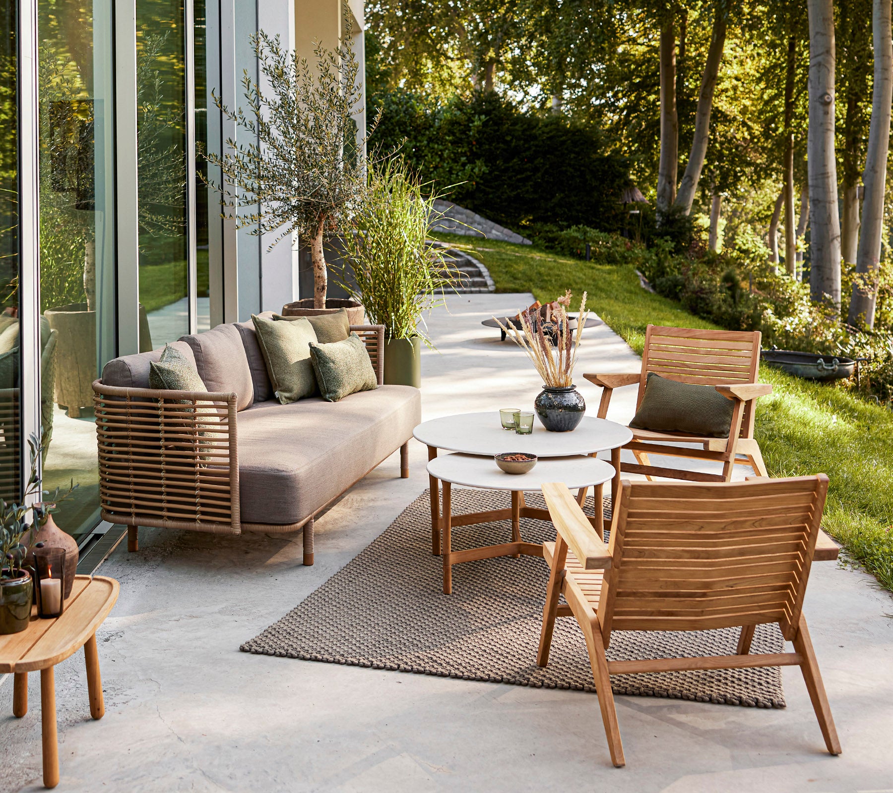 Stilvoller Außenbereich mit einem modernen Sofa, Holzstühlen, einem runden Tisch und dekorativen Pflanzen vor einer ruhigen Kulisse.