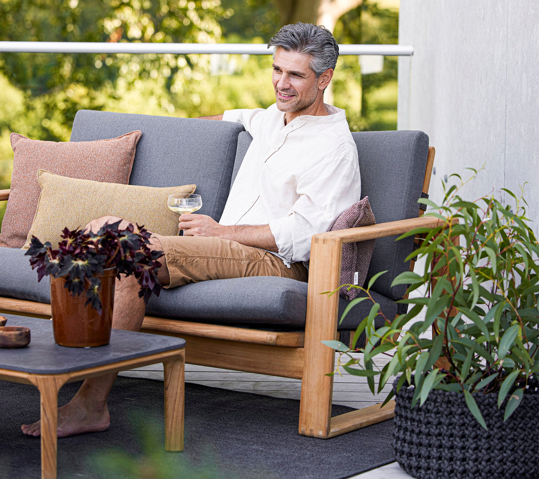 Ein Mann genießt ein Getränk, während er auf einem bequemen Sofa sitzt, umgeben von Pflanzen und dekorativen Kissen.