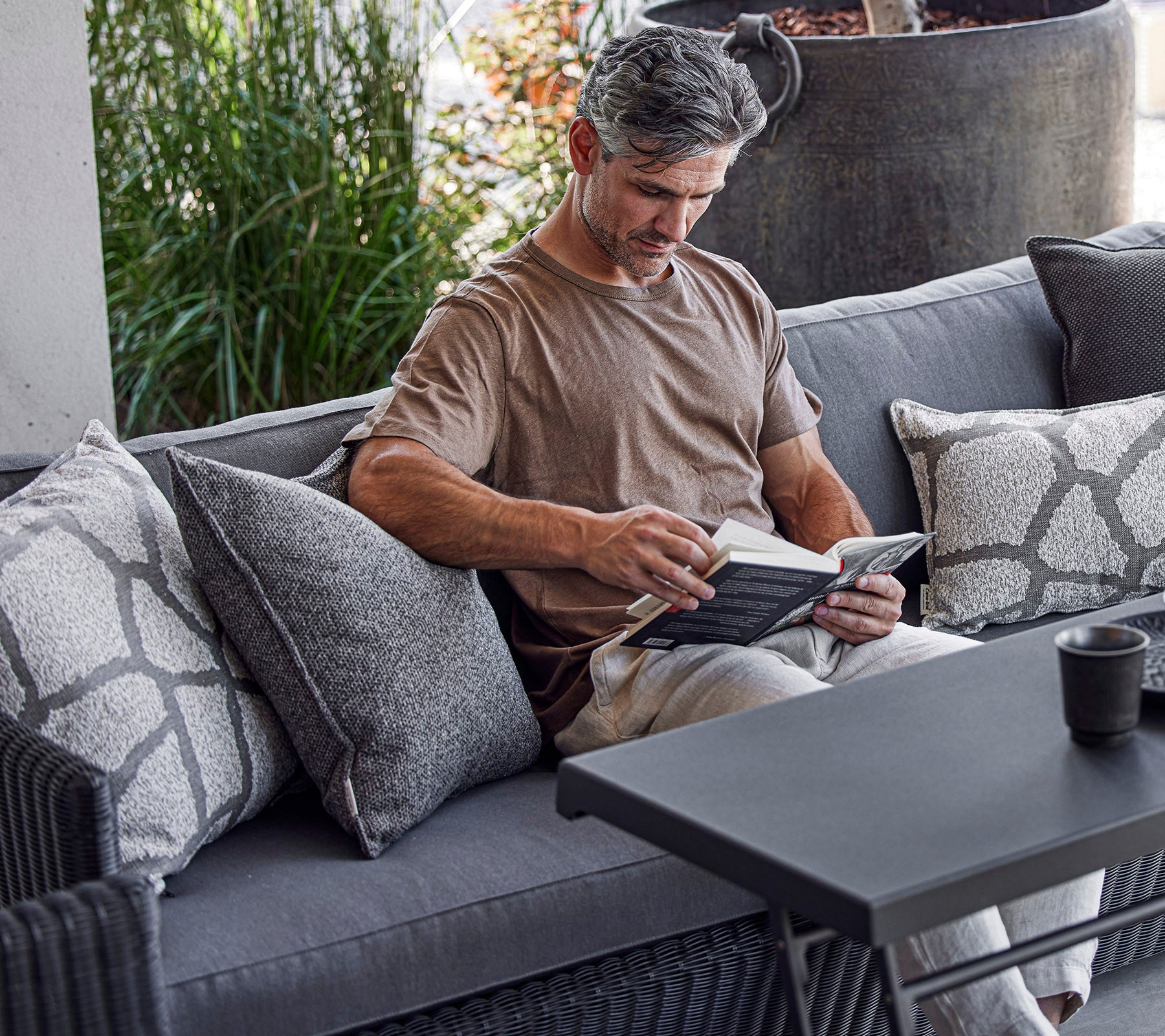 Ein Mann liest ein Buch, während er auf einem gemütlichen Sofa mit dekorativen Kissen sitzt, mit Grünpflanzen im Hintergrund.