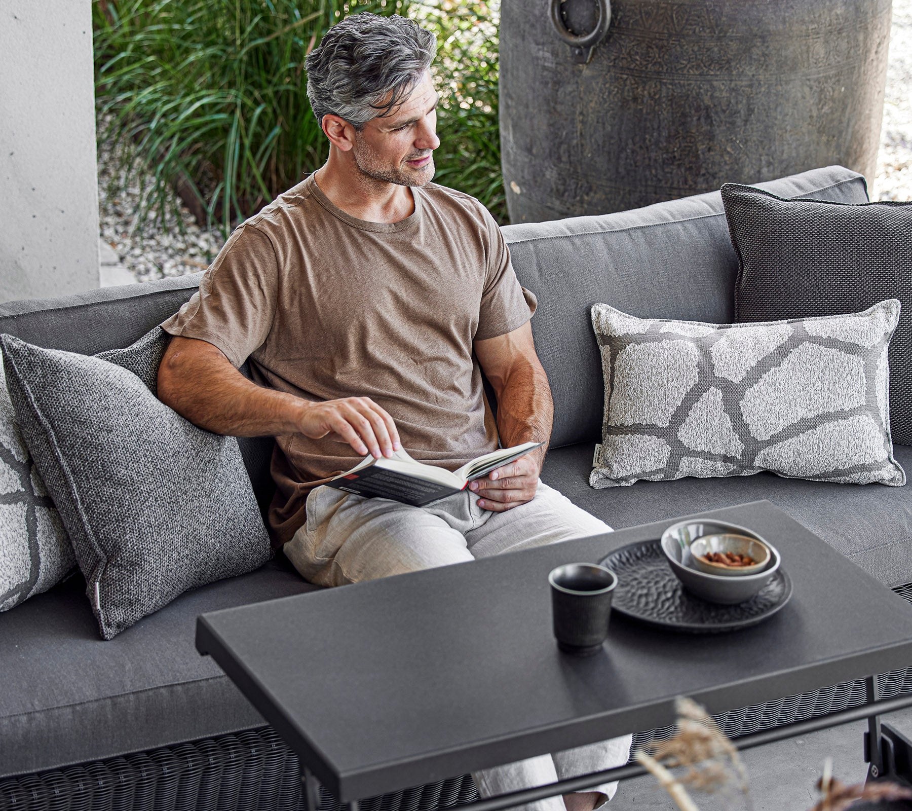 Ein Mann sitzt bequem auf einem Sofa, liest ein Buch mit einer Tasse und einer Schüssel auf einem Couchtisch neben ihm, umgeben von dekorativen Kissen.