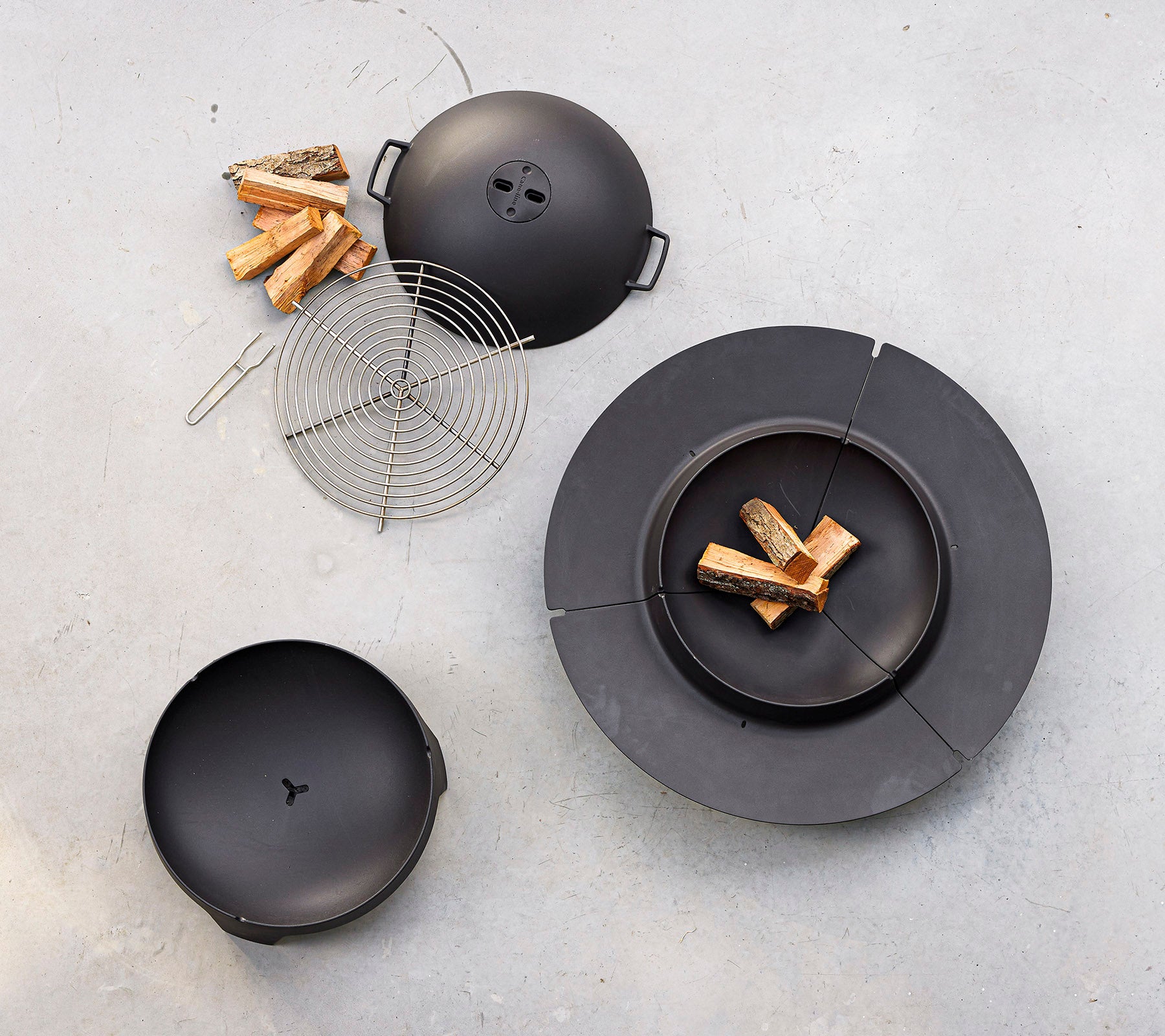 Ein schwarzes Kochset mit einem runden Grill mit Deckel, einem Grillrost und Holzstücken, die in der Nähe angeordnet sind.