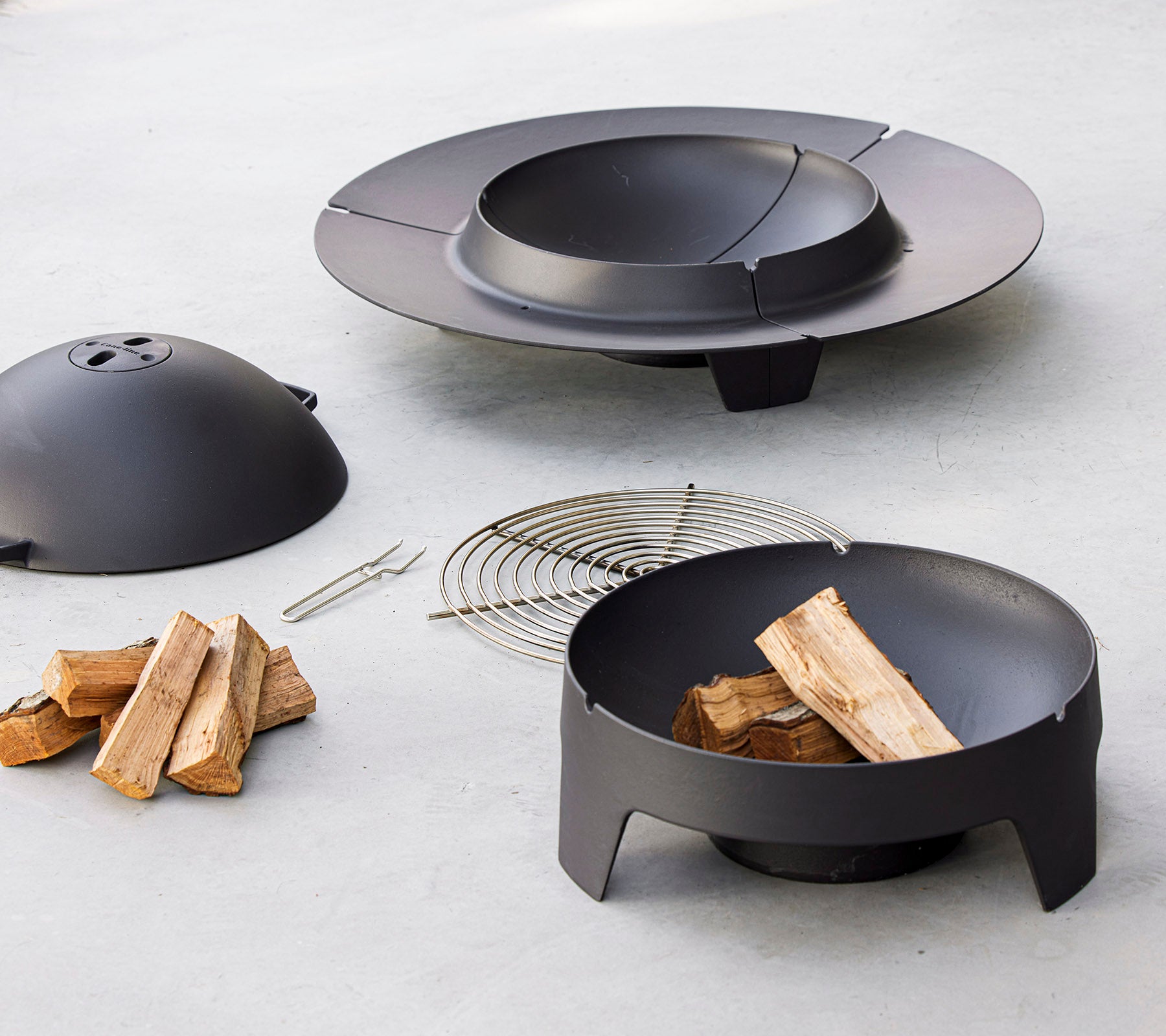 Ein schwarzes Feuerschalen-Set mit einer Schüssel, einem Deckel, einem Rost und gestapeltem Holz, gestaltet für Außenveranstaltungen.